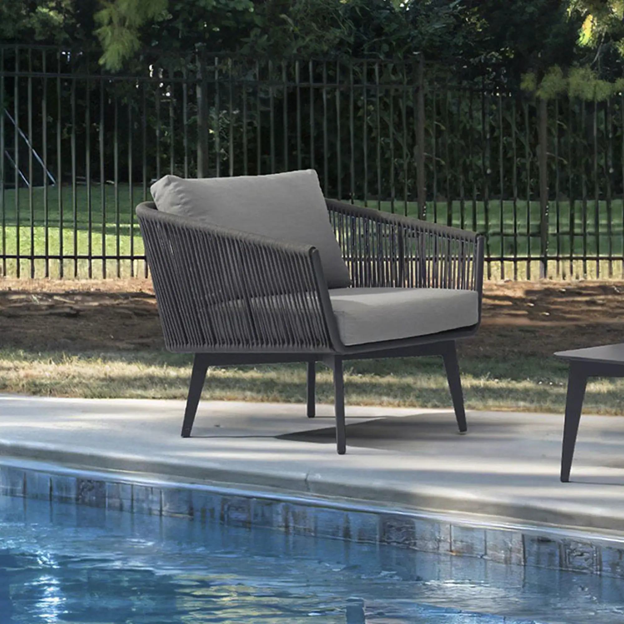 A modern dark gray woven outdoor armchair with light gray cushions sits on a concrete patio next to a blue swimming pool. A dark fence and green trees are in the background.