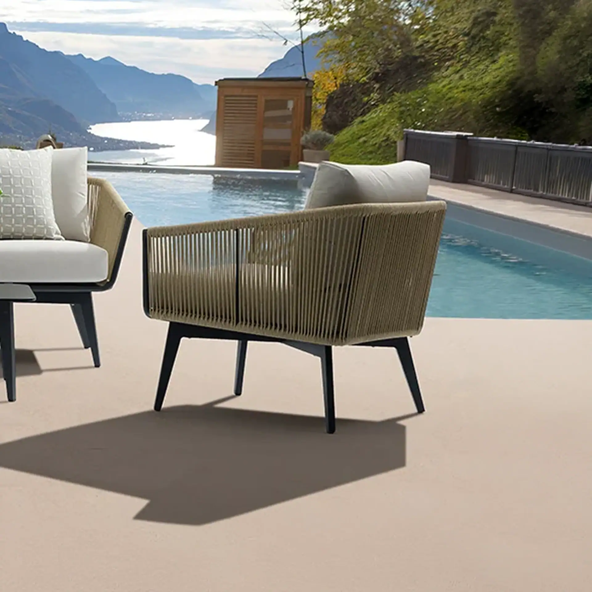 Two modern outdoor armchairs with woven backs and white cushions sit on a patio next to an infinity pool, overlooking a scenic mountain lake.
