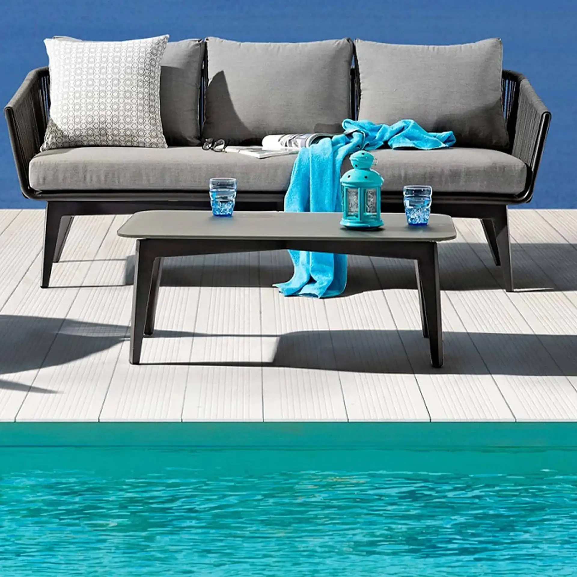 Outdoor grey sofa and coffee table on a light wooden deck next to a turquoise swimming pool. The sofa has cushions, a patterned pillow, a blue towel, and sunglasses. The table holds two glasses of water and a turquoise lantern.