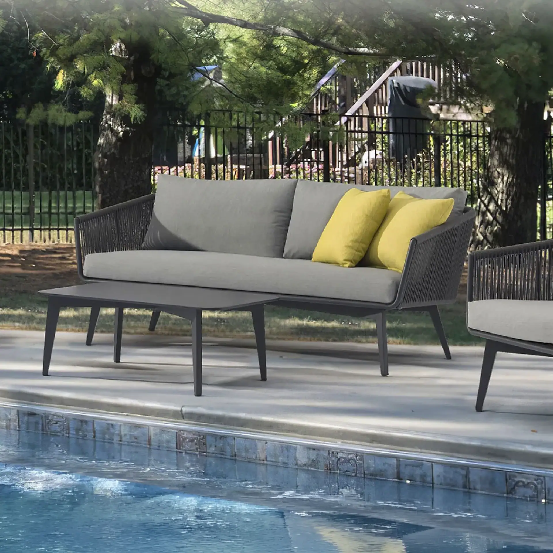 A gray outdoor sofa with yellow pillows and a dark gray coffee table next to a swimming pool, with a matching chair partially visible. Trees and a fence are in the background.