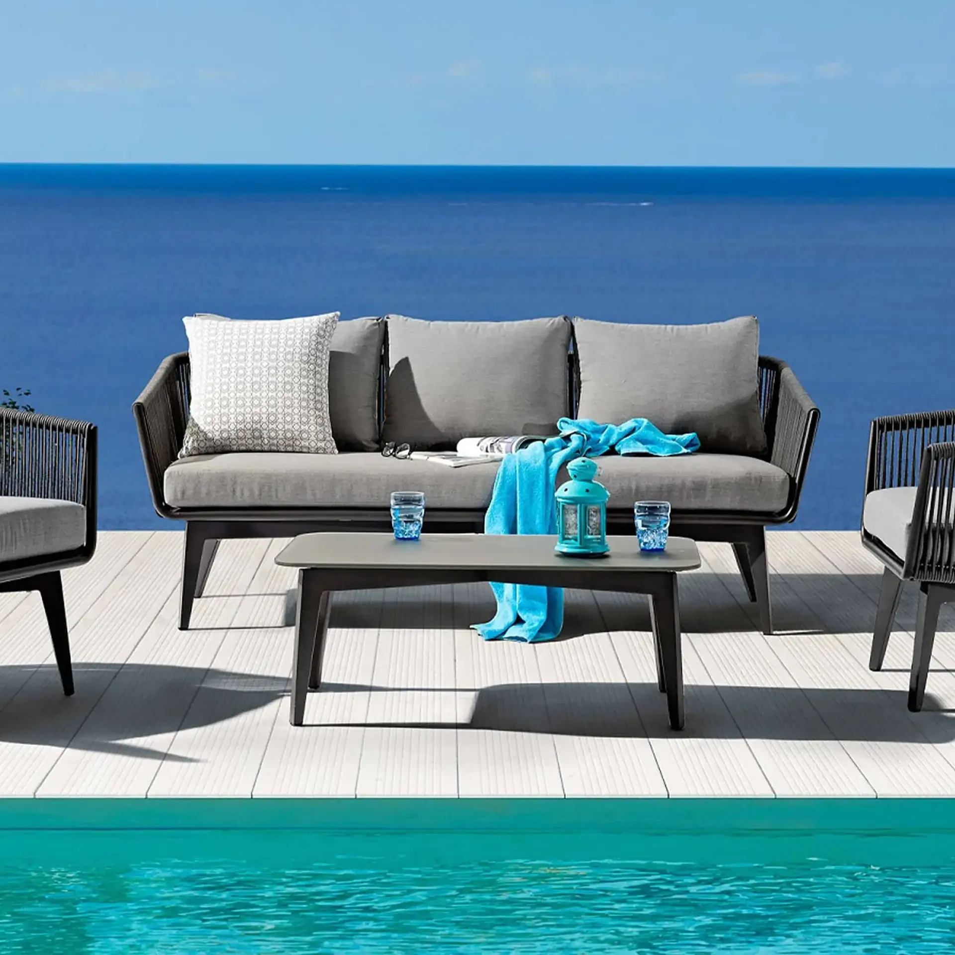 An outdoor lounge area with gray sofa and armchairs on a light wooden deck, overlooking a blue ocean and a turquoise swimming pool.