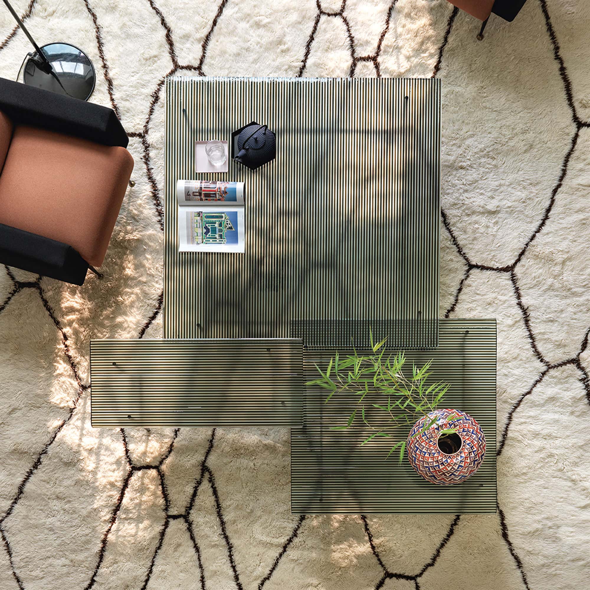 A glass coffee table with magazines, a decorative vase, and a plant, set on a patterned rug in a stylish living room.