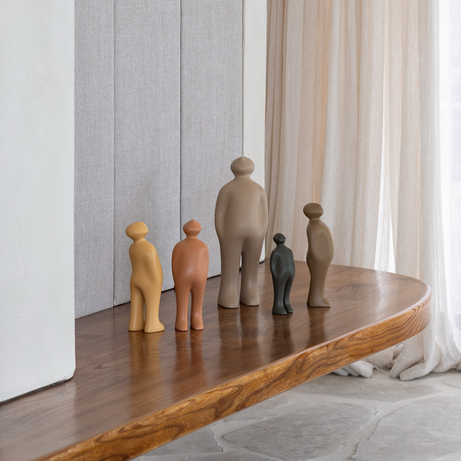 Five abstract, human-like figurines in varying sizes and earthy colors (yellow, terracotta, beige, dark grey, light brown) lined up on a polished wooden surface. A grey textured wall and sheer curtains are in the background.