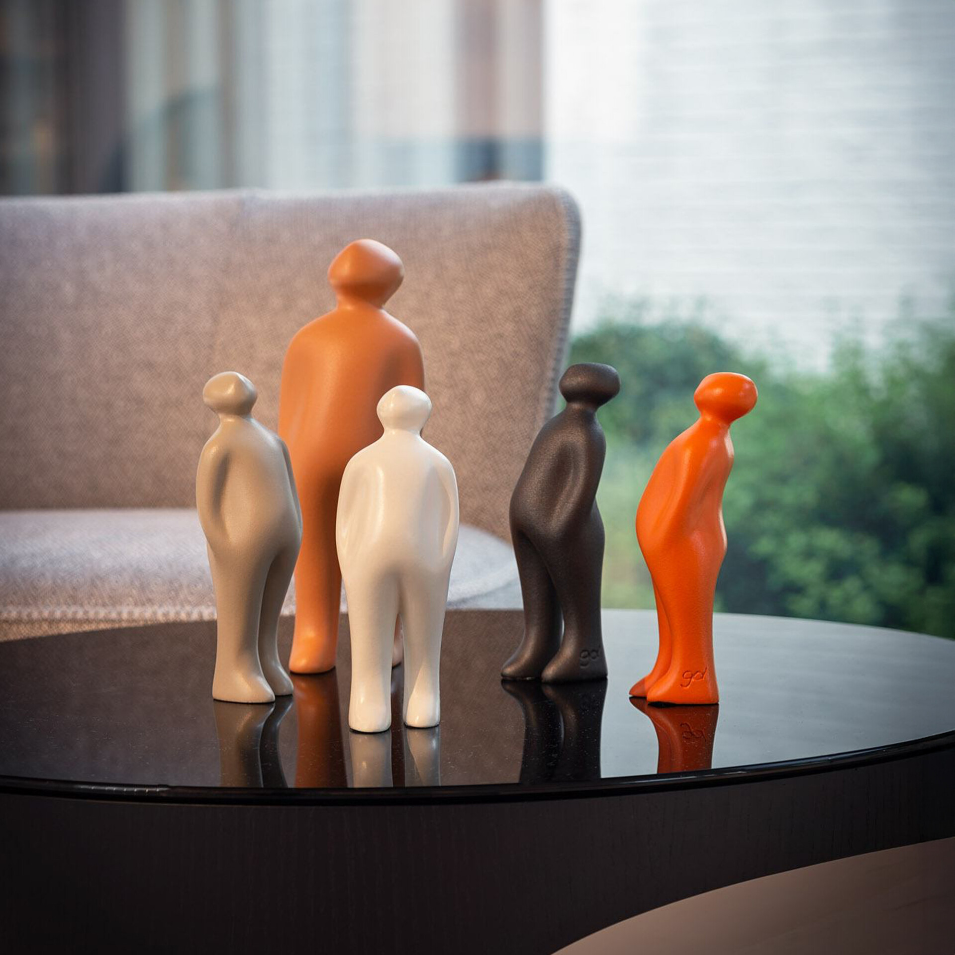 Five abstract human-like figurines in various colors (light gray, white, dark gray, bright orange, and burnt orange) stand on a glossy black table. Some figures are leaning. Their reflections are visible on the table. A blurred light-colored armchair and a window overlooking green foliage are in the background.