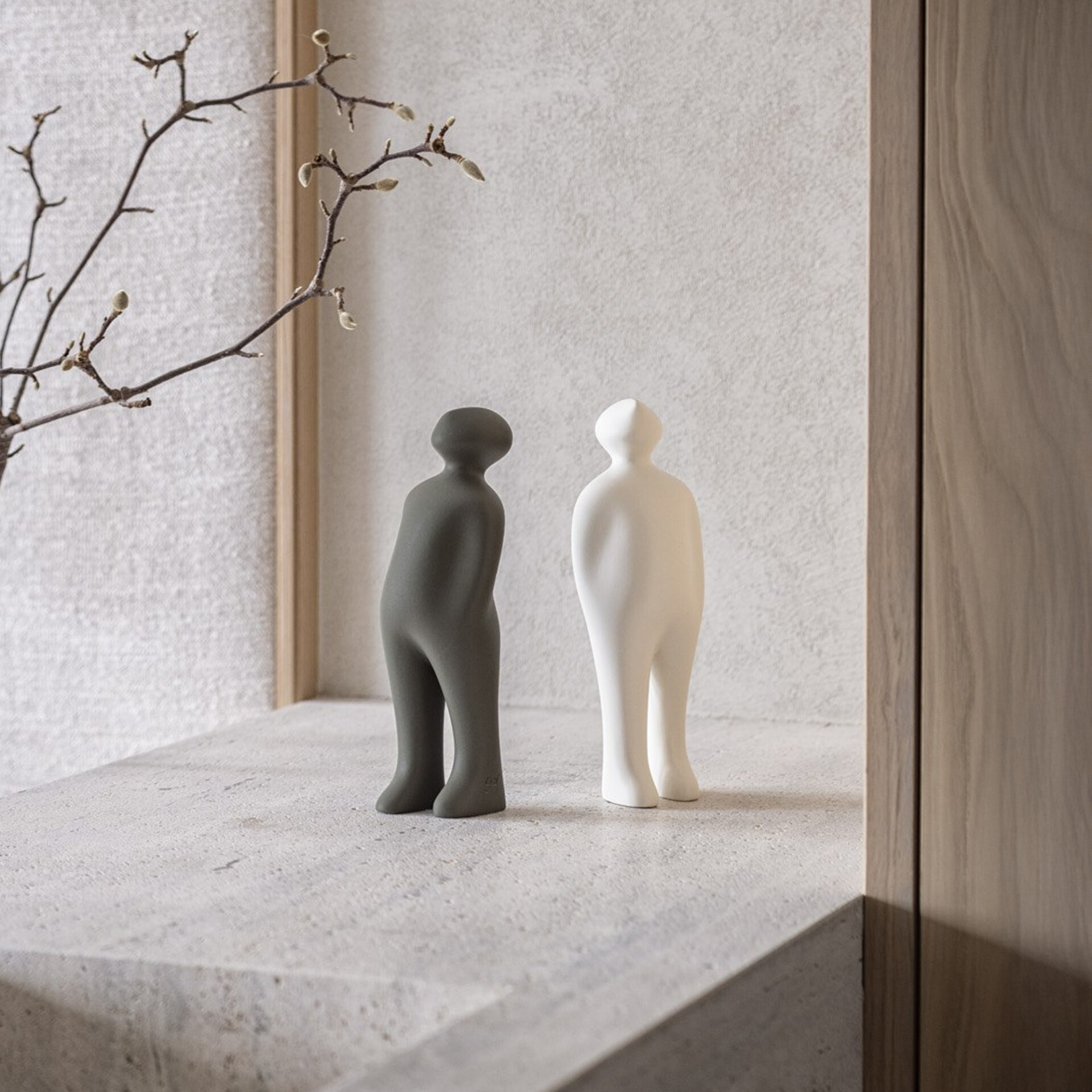 Two minimalist human-like figurines, one dark grey and one off-white, stand on a light grey textured shelf. Bare branches with buds are blurred in the background to the left, and a wooden panel is visible to the right.