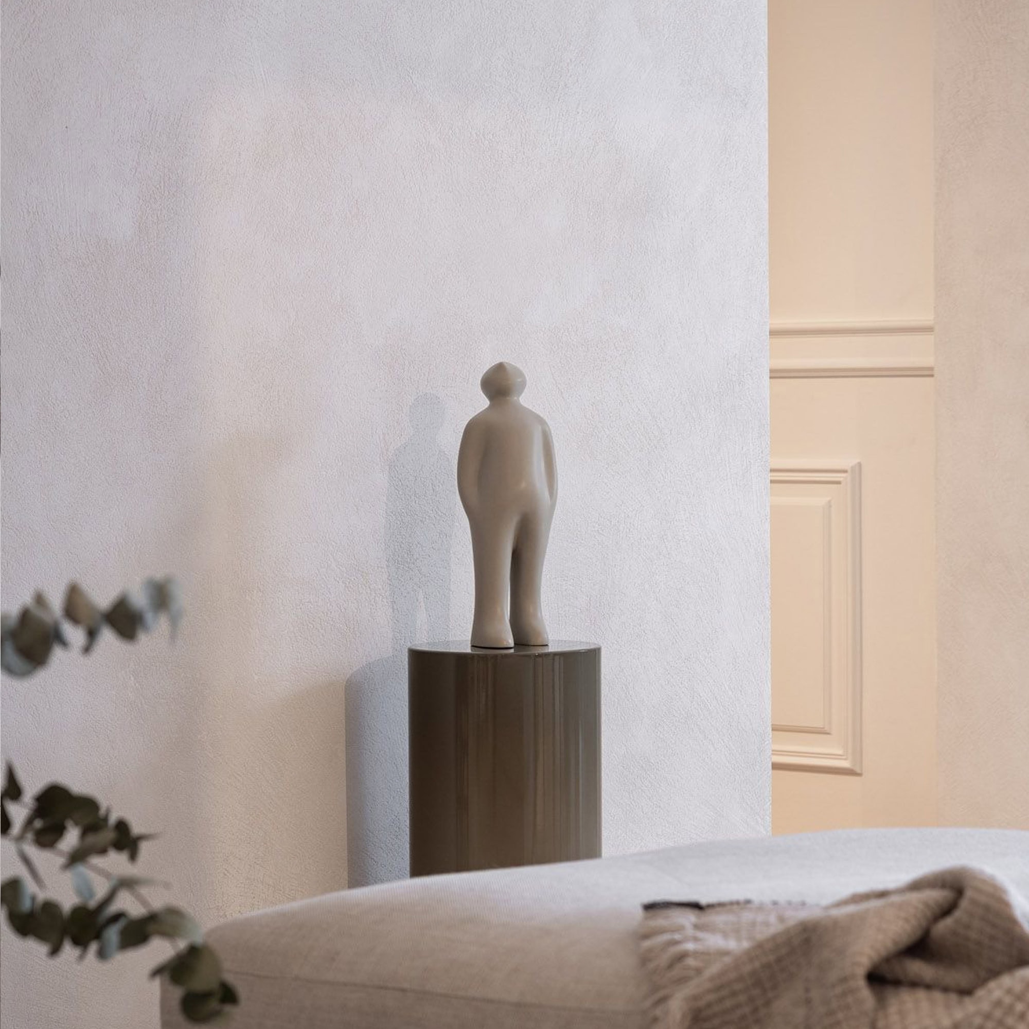 A light grey abstract human-like sculpture on a dark grey cylindrical pedestal, positioned in front of a textured off-white wall. A blurred beige upholstered furniture piece with a throw blanket and eucalyptus leaves are in the foreground.
