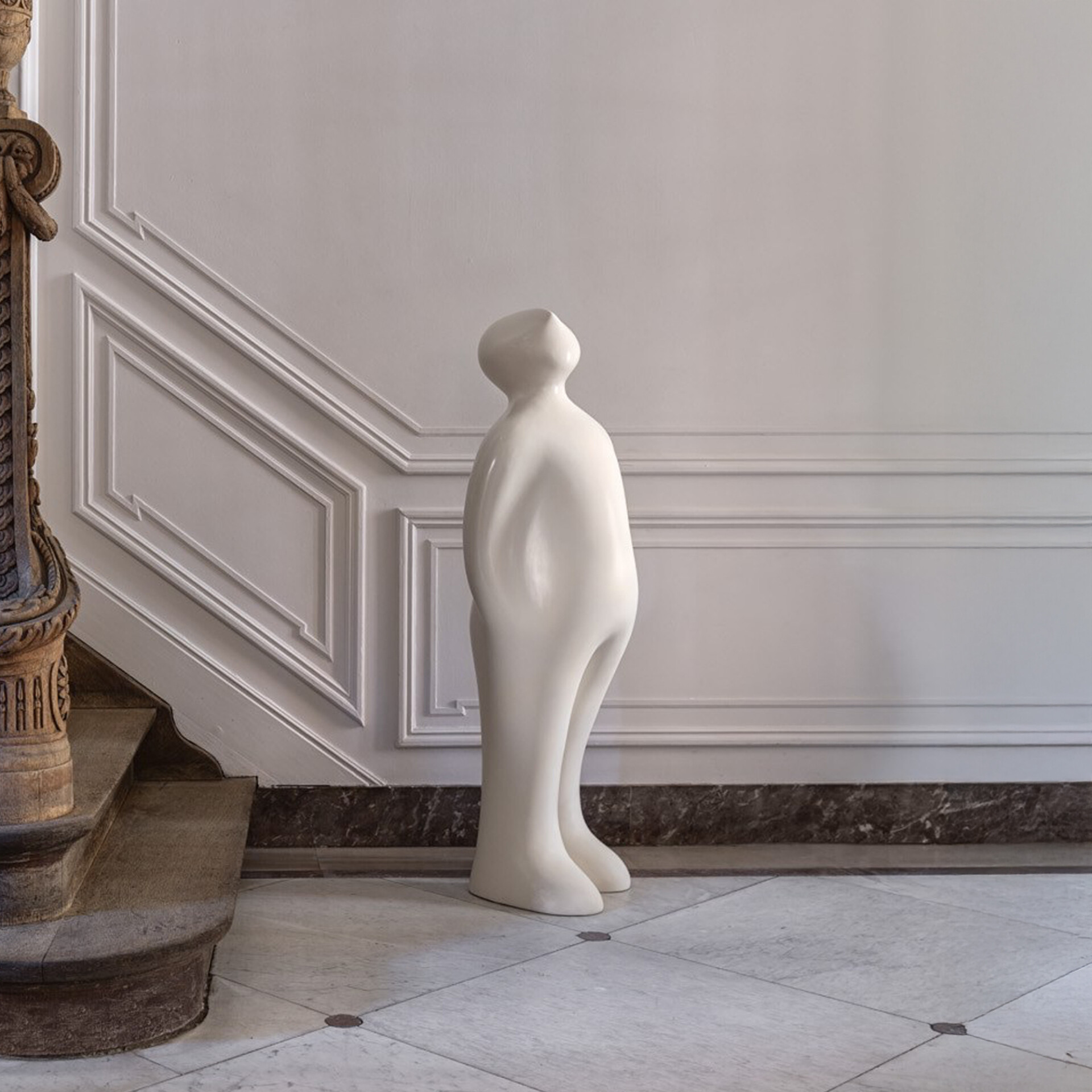 A tall, smooth, white abstract human-like sculpture stands in a grand hall with intricate white wall paneling, a marble floor, and a carved wooden staircase.