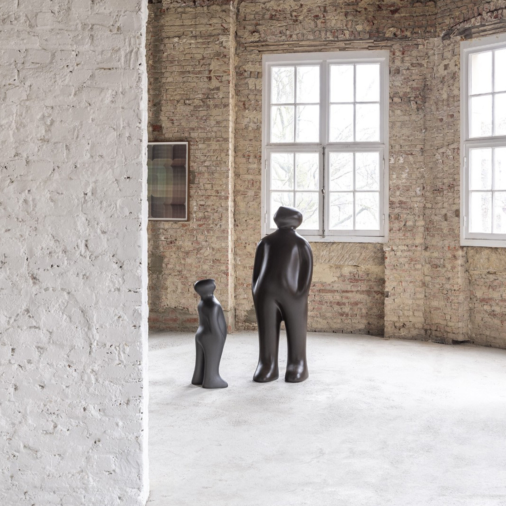 Two dark, abstract human-like sculptures stand in a room with exposed brick walls and large windows. A larger figure faces away, and a smaller figure looks up at it.