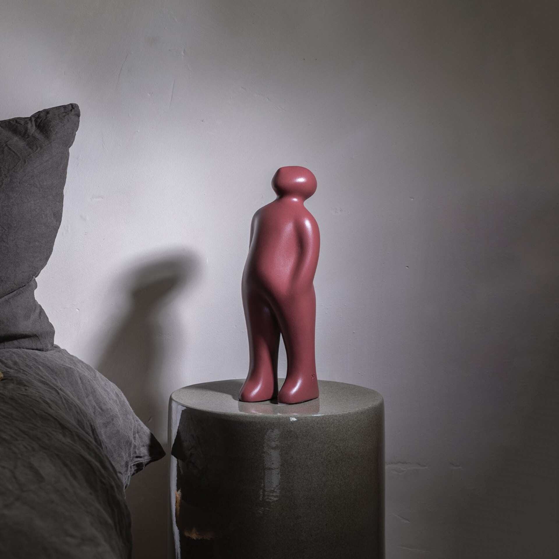 A smooth, muted rose abstract human-like sculpture stands on a dark gray cylindrical pedestal against a light wall. A long shadow of the sculpture is cast behind it, and dark gray textured fabric is visible in the lower-left foreground.