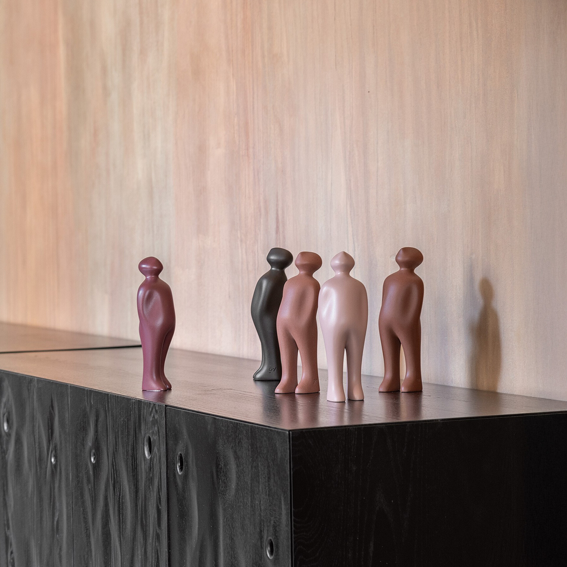 Five abstract, human-like sculptures in various colors (purple, black, terracotta, pink, brown) stand on a dark wooden cabinet against a light wood wall.