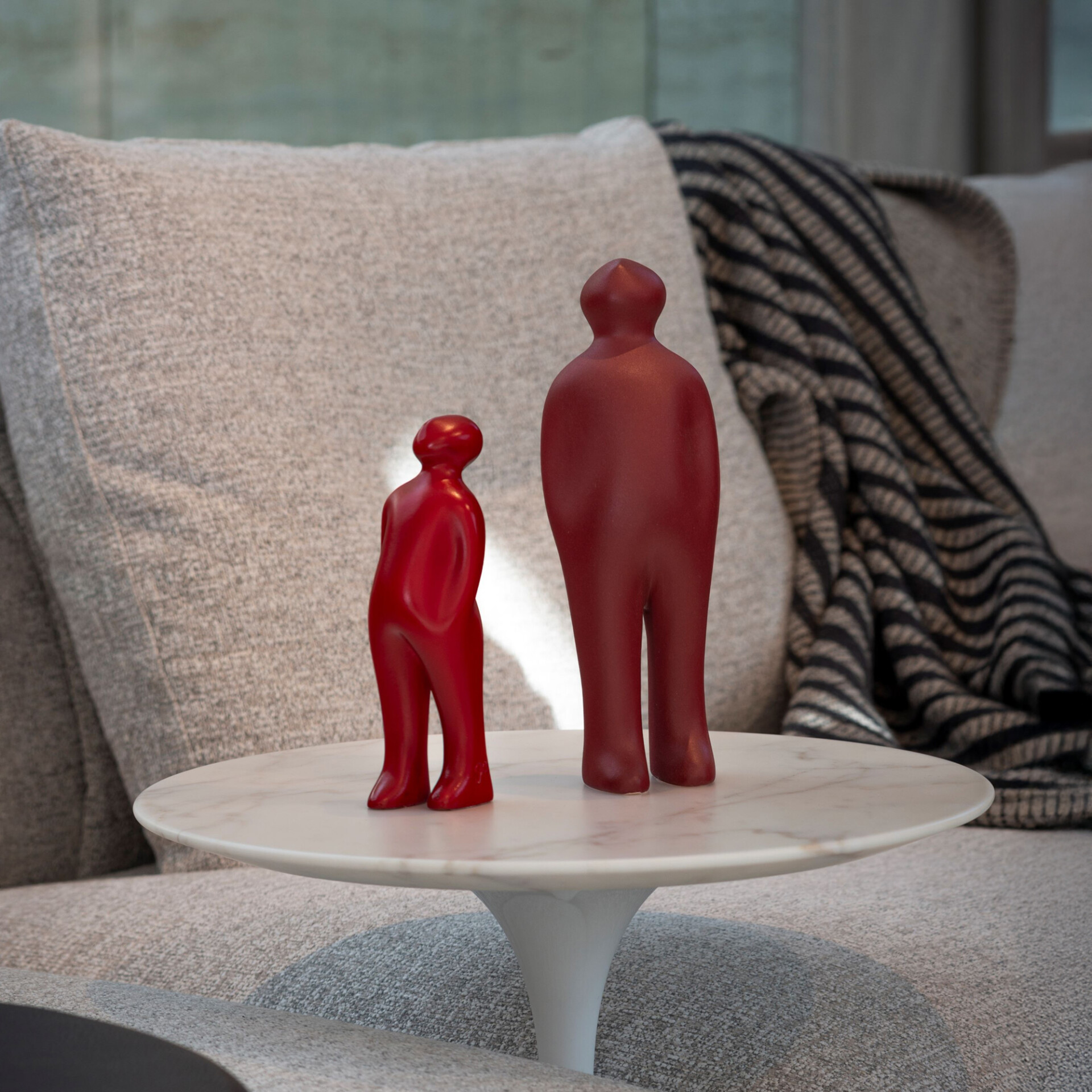 Two matte red abstract human-like figurines, one taller and one shorter, stand on a round white marble side table in front of a light gray sofa with a striped throw blanket.