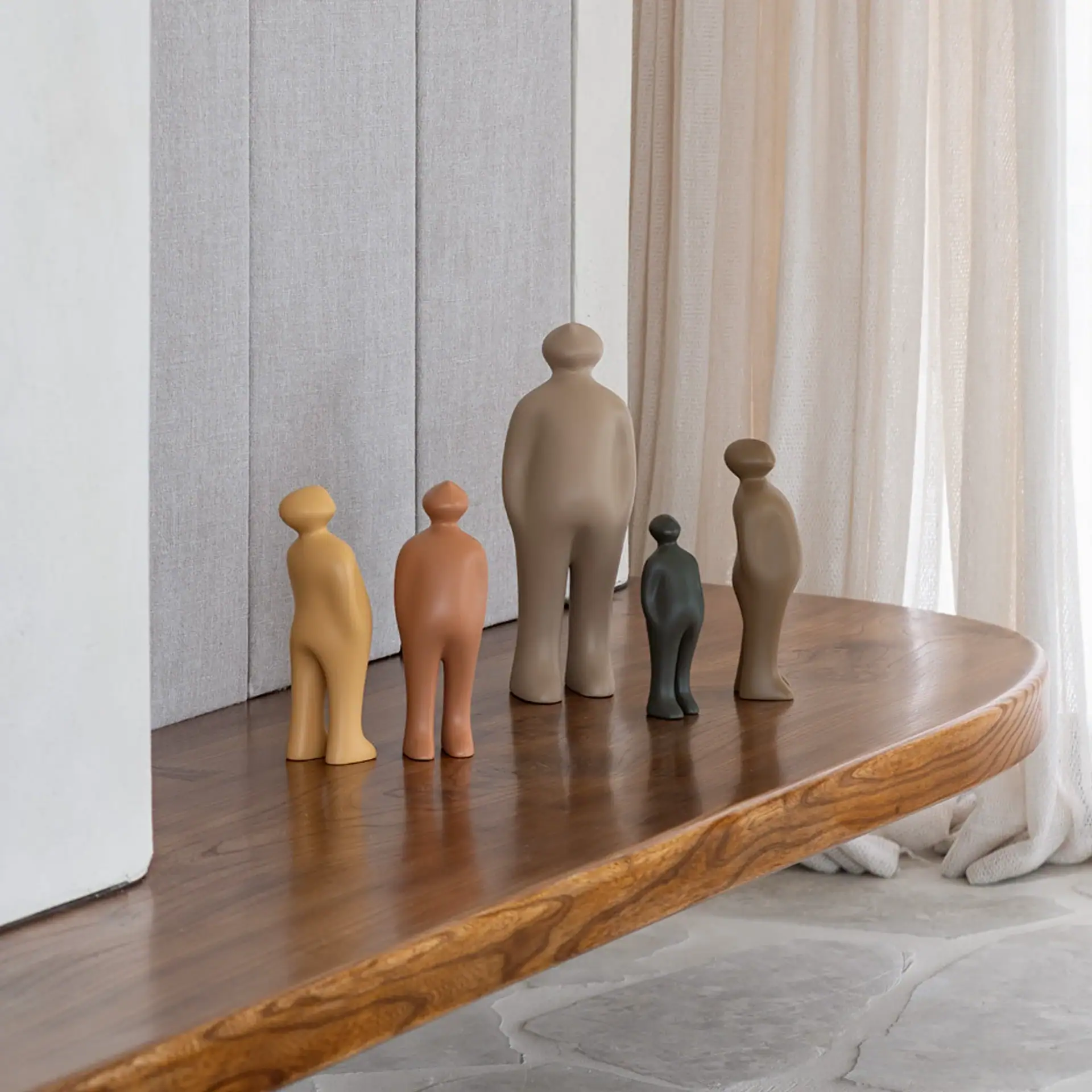 A light grey abstract human-like sculpture on a dark grey cylindrical pedestal, positioned in front of a textured off-white wall. A blurred beige upholstered furniture piece with a throw blanket and eucalyptus leaves are in the foreground.