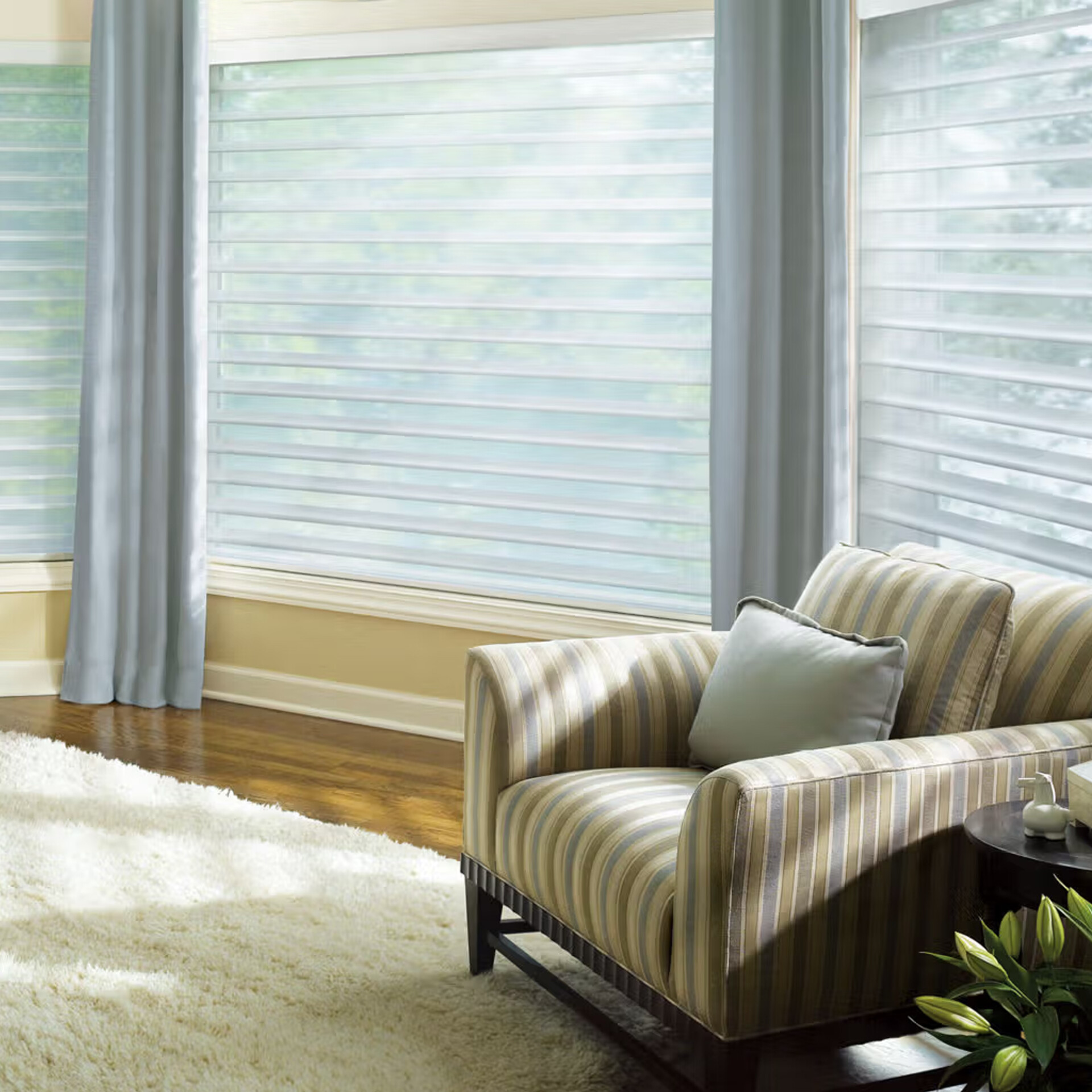 A cozy living room corner with large windows covered by sheer horizontal blinds and light blue drapes. A striped armchair, a plush cream rug, and a dark side table with lilies are visible.