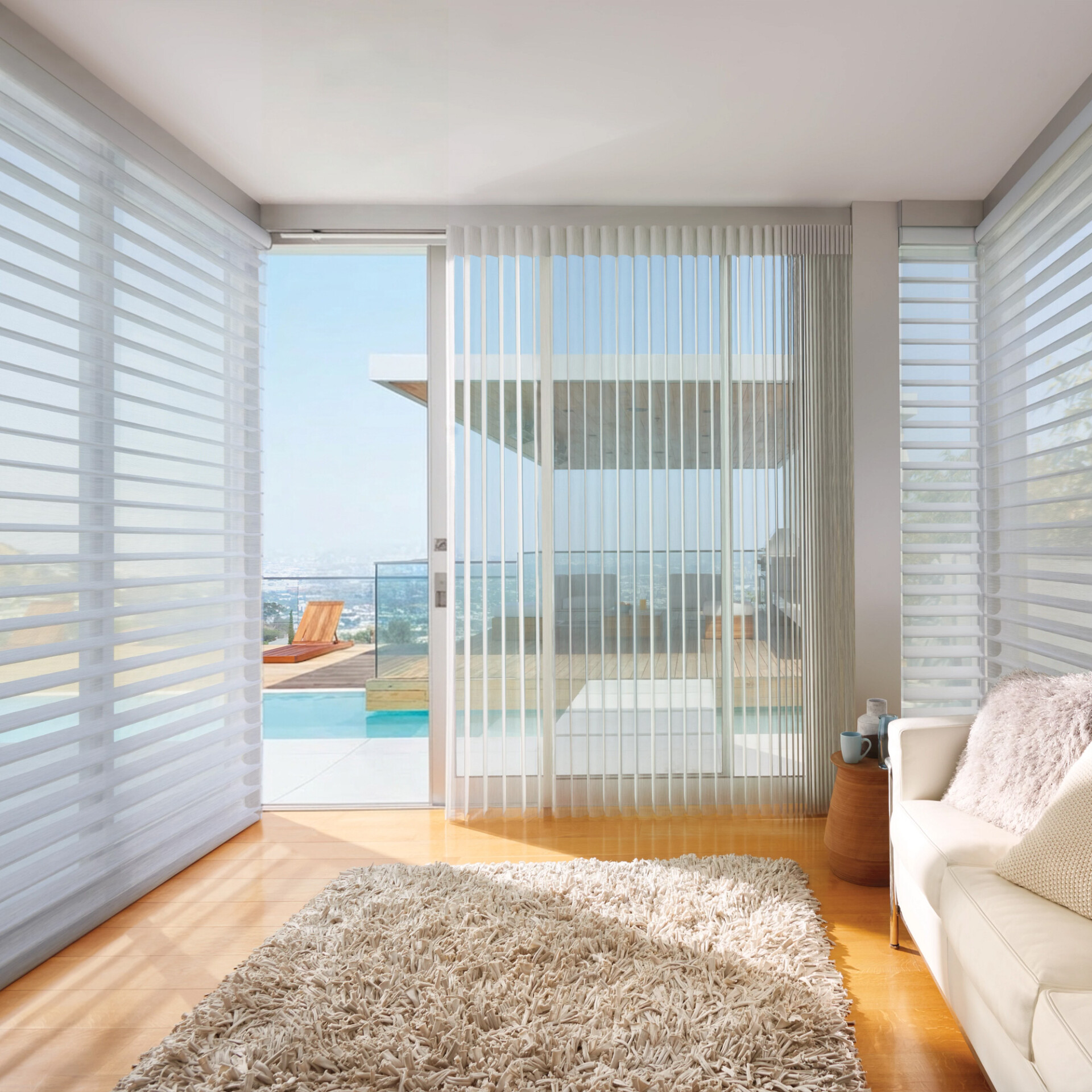 A modern living room with light hardwood floors, a shaggy beige rug, and a white leather sofa. Large windows and a sliding glass door with sheer white blinds overlook a patio with a blue swimming pool and a panoramic city view under a clear sky.