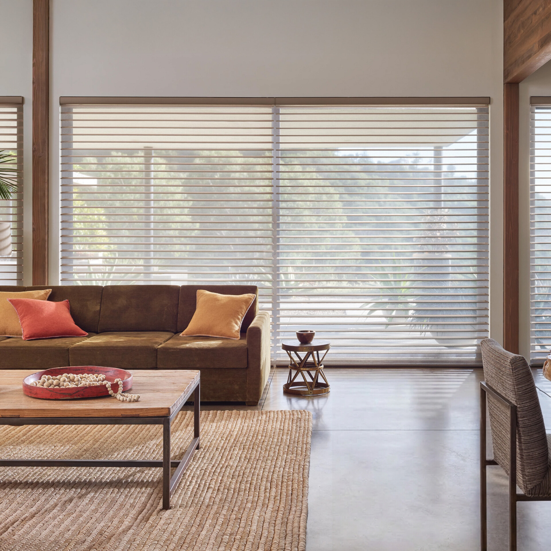 A modern living room with a brown sectional sofa, wooden coffee table on a woven rug, and large windows with sheer horizontal shades diffusing natural light. Greenery is visible outside.