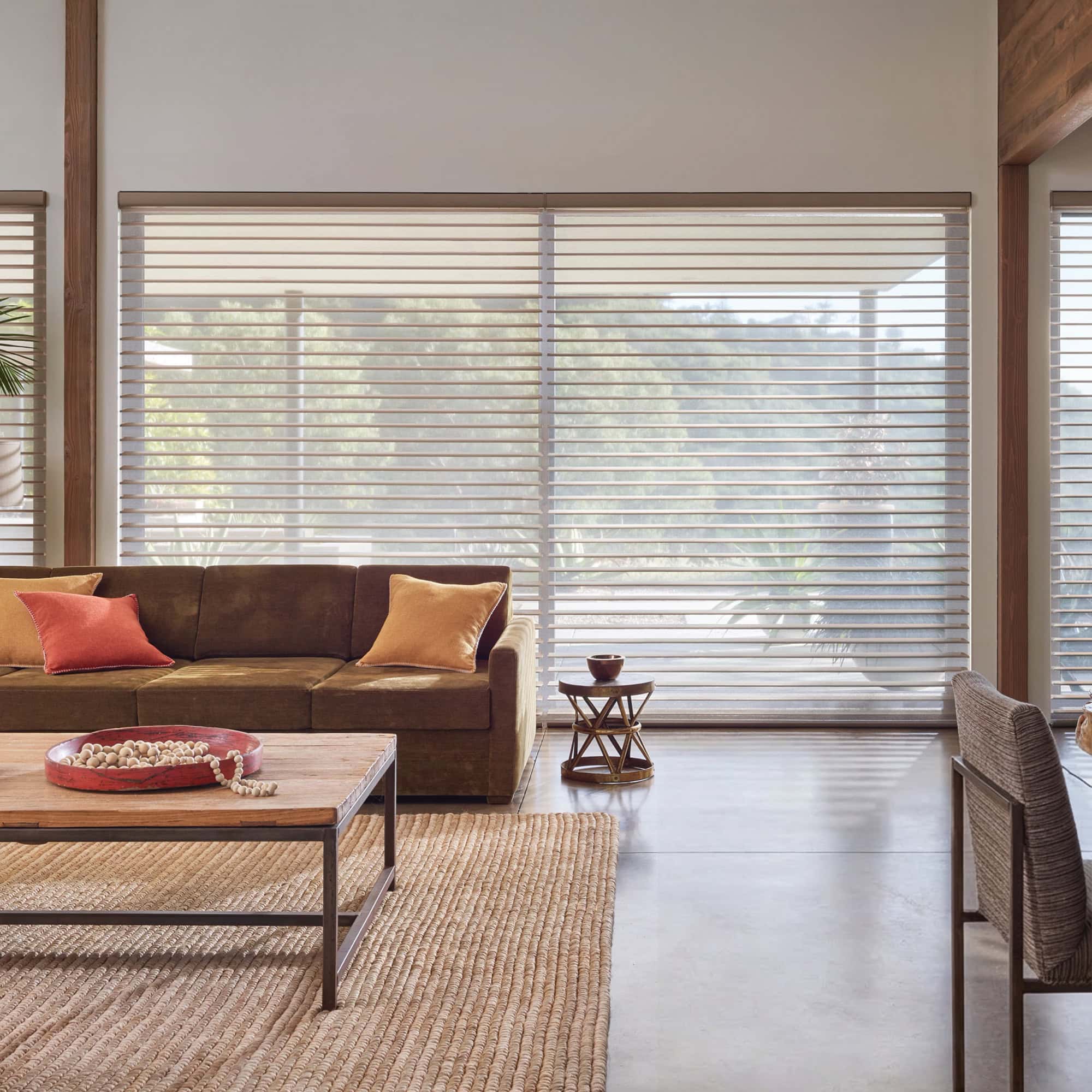A modern living room with a brown sectional sofa, wooden coffee table on a woven rug, and large windows with sheer horizontal shades diffusing natural light. Greenery is visible outside.