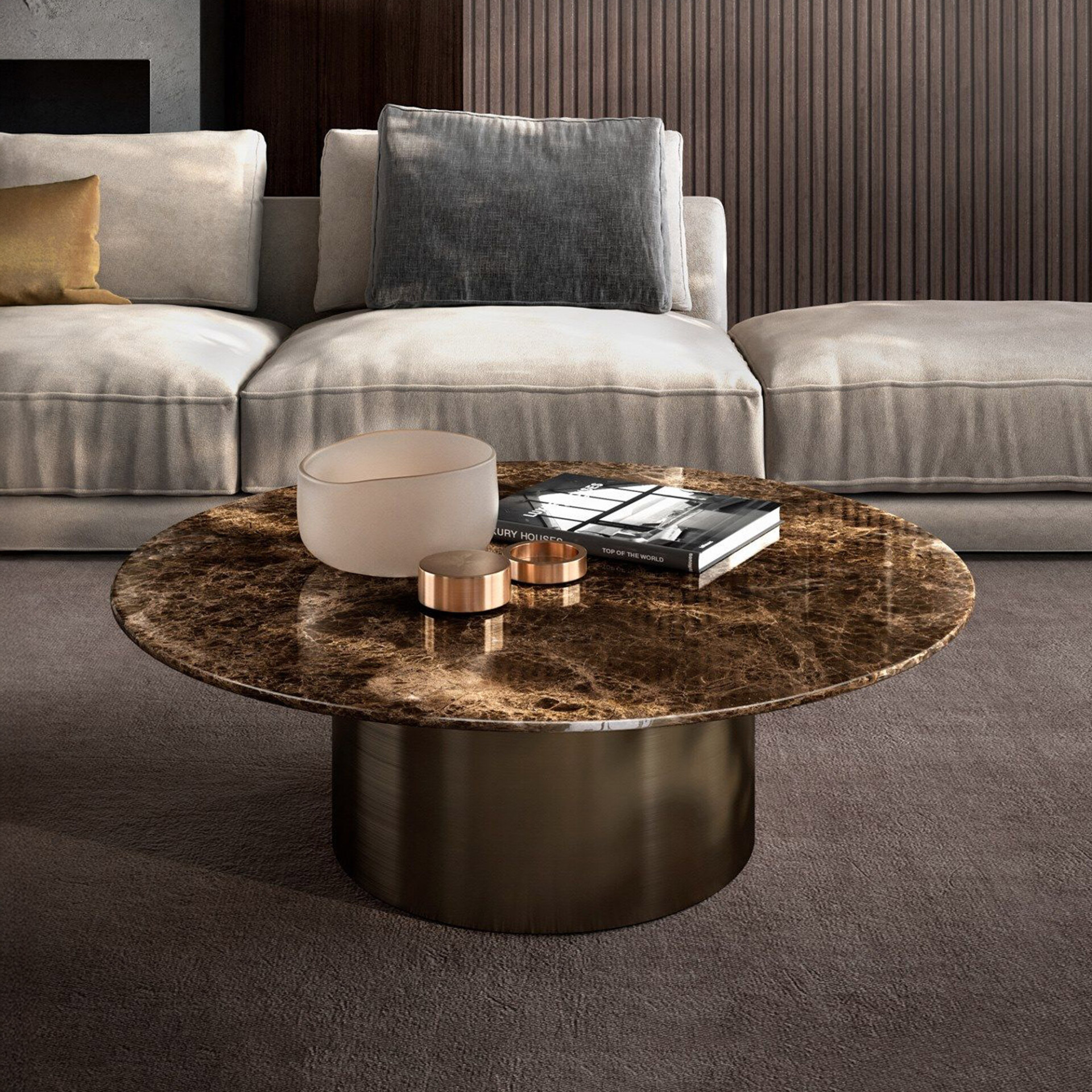 A luxurious living room with a round, dark brown marble coffee table on a bronze base. The table holds a frosted bowl, metallic rings, and a 'Luxury Houses' book. Behind it is a light-colored sofa with a golden and a grey pillow, against a dark wood-paneled wall and dark carpet.