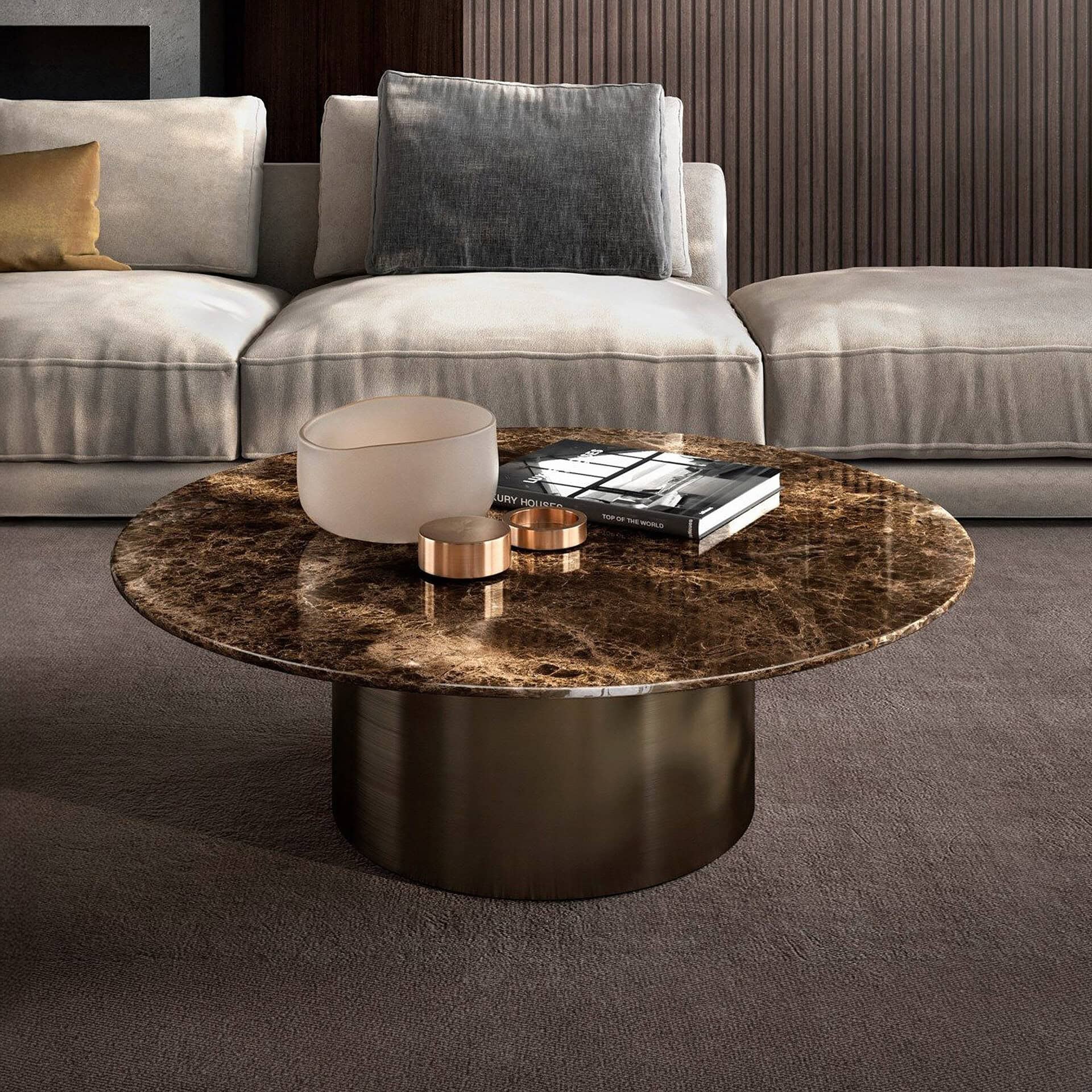 A luxurious living room with a round, dark brown marble coffee table on a bronze base. The table holds a frosted bowl, metallic rings, and a 'Luxury Houses' book. Behind it is a light-colored sofa with a golden and a grey pillow, against a dark wood-paneled wall and dark carpet.