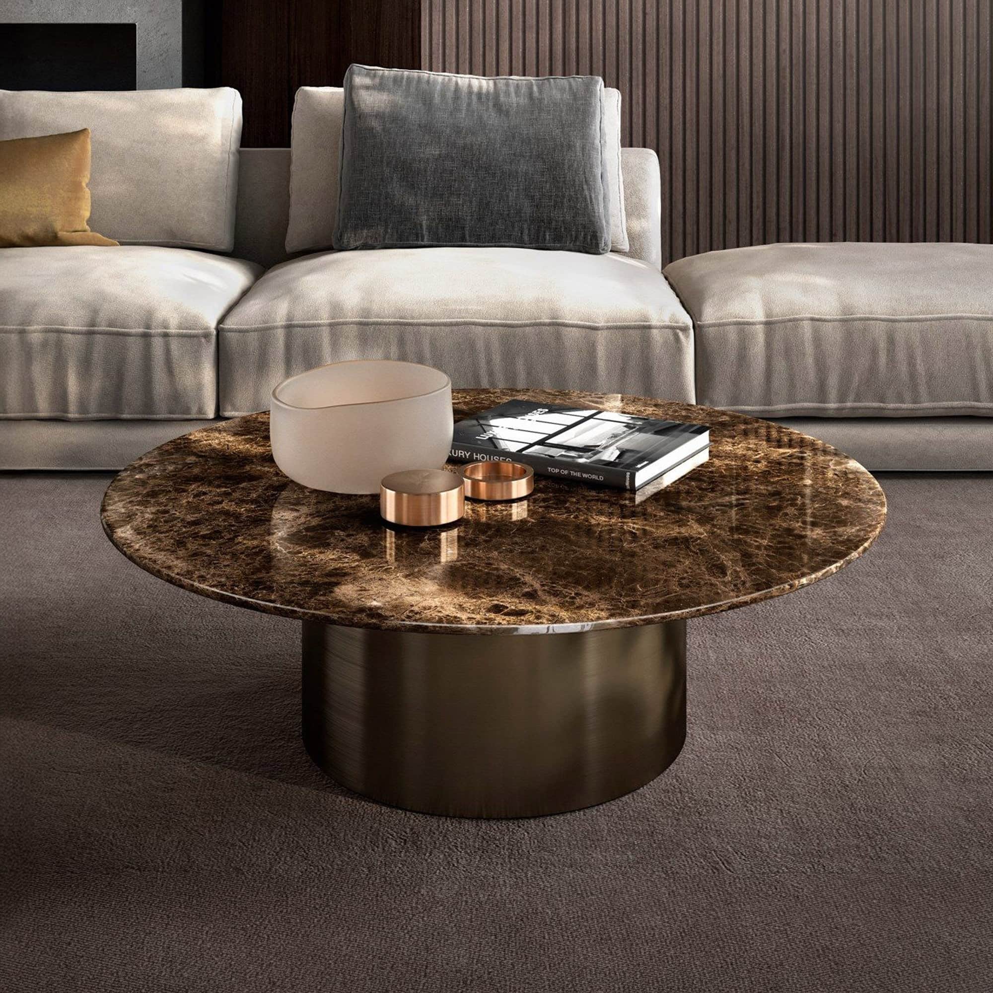 A luxurious living room with a round, dark brown marble coffee table on a bronze base. The table holds a frosted bowl, metallic rings, and a 'Luxury Houses' book. Behind it is a light-colored sofa with a golden and a grey pillow, against a dark wood-paneled wall and dark carpet.
