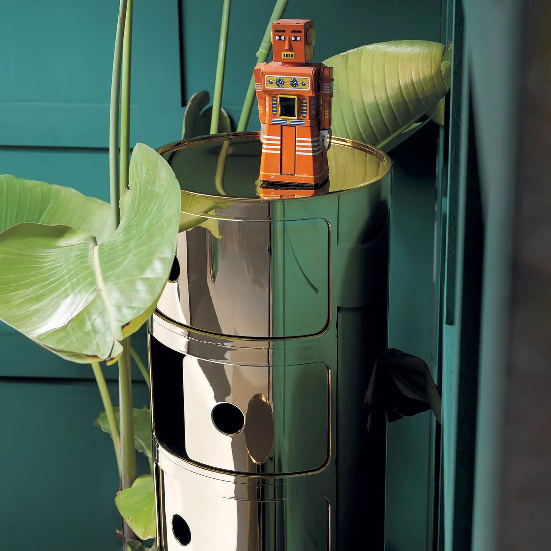 Modern robot figurine on metallic planter with tropical leaves.