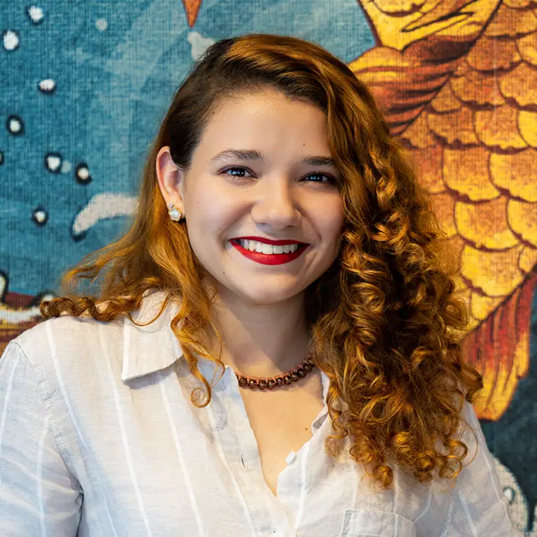 A young woman with curly reddish-brown hair and red lipstick smiles, wearing a striped shirt and pearl earrings, against a colorful tapestry with a fish design.