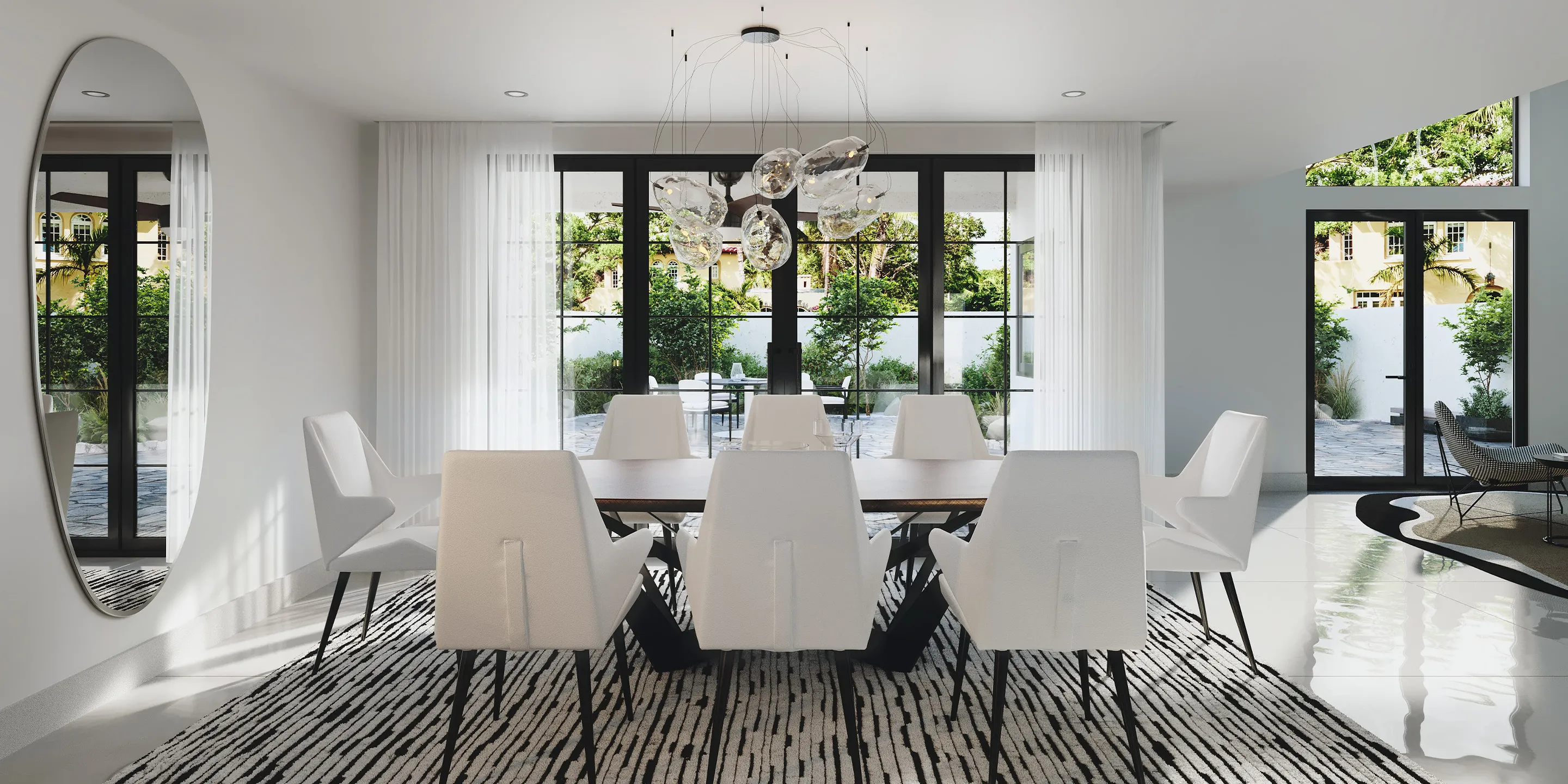 A modern dining room with a long wooden table, eight white upholstered chairs, and a unique glass chandelier. Large black-framed windows overlook a lush green garden and patio.