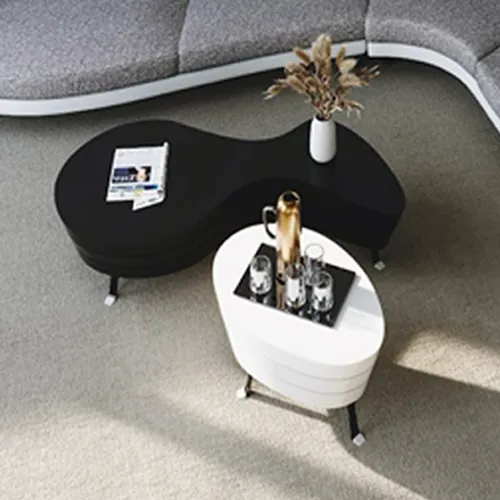 High-angle view of a modern living room with a curved gray sectional sofa, a black organic-shaped coffee table with a magazine and vase, and a white oval coffee table with a gold carafe and glasses.
