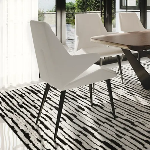 A modern dining room with a white angular chair on a black and white patterned rug, a wooden table, and a large window looking out to greenery.