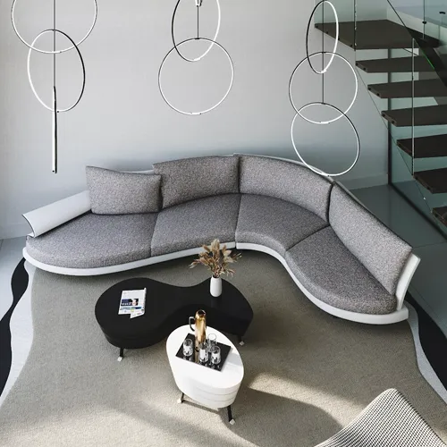 A high-angle view of a modern living room with a curved gray sectional sofa, two organic-shaped coffee tables, and circular pendant lights. A glass-railed staircase is visible in the background.