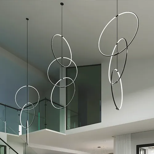 Three modern pendant light fixtures, each made of multiple interconnected illuminated rings, hang from a high ceiling in a contemporary interior with glass railings.