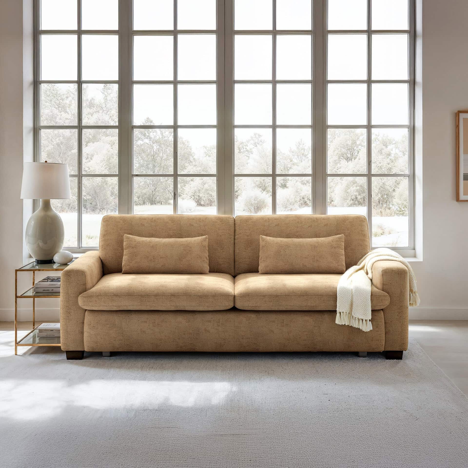 A tan sofa with a cream throw blanket sits in a bright living room in front of a large, multi-pane window overlooking a snowy winter landscape. A lamp is on a gold side table to the left.
