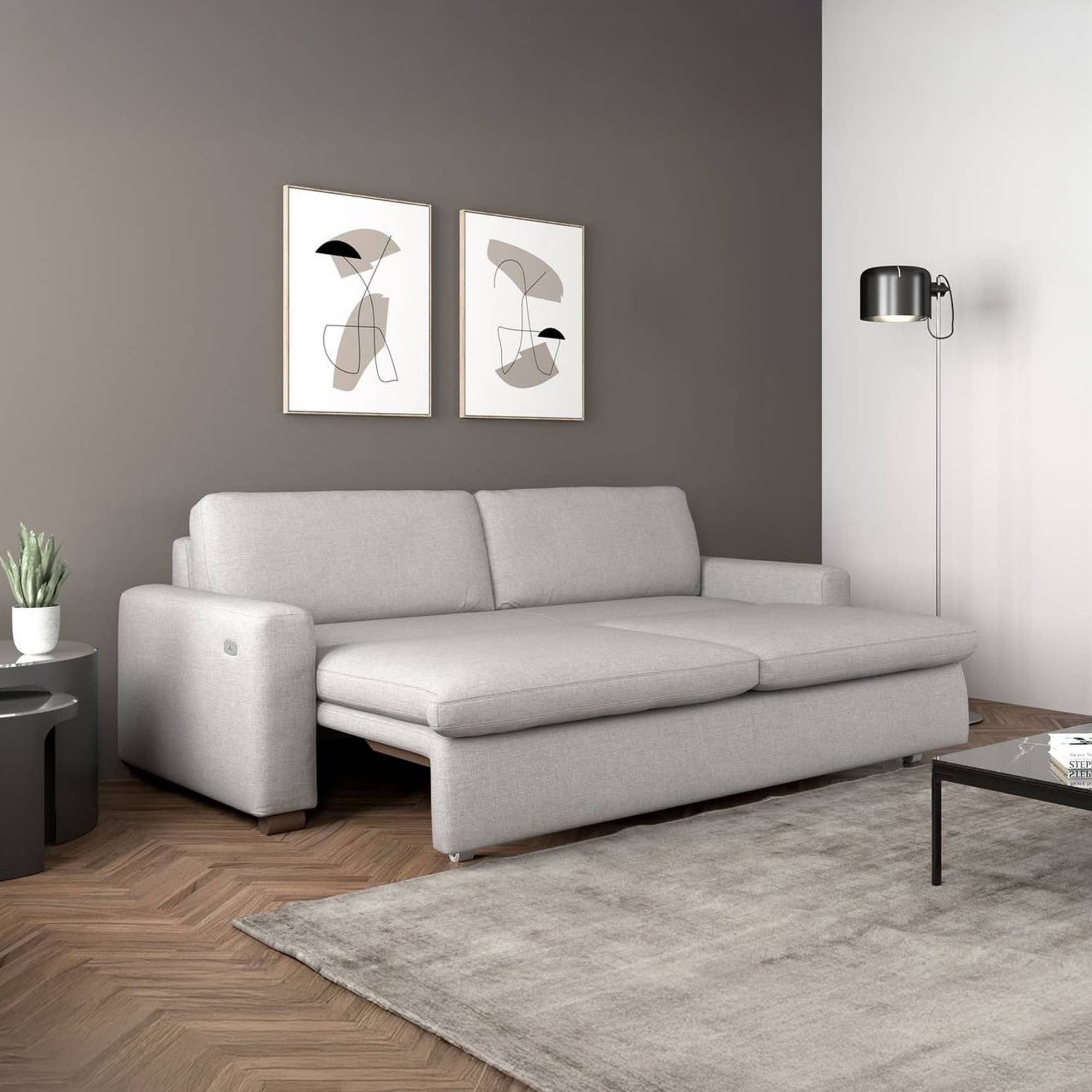 A light grey pull-out sofa in a modern living room with abstract art, a black floor lamp, and a herringbone wood floor.