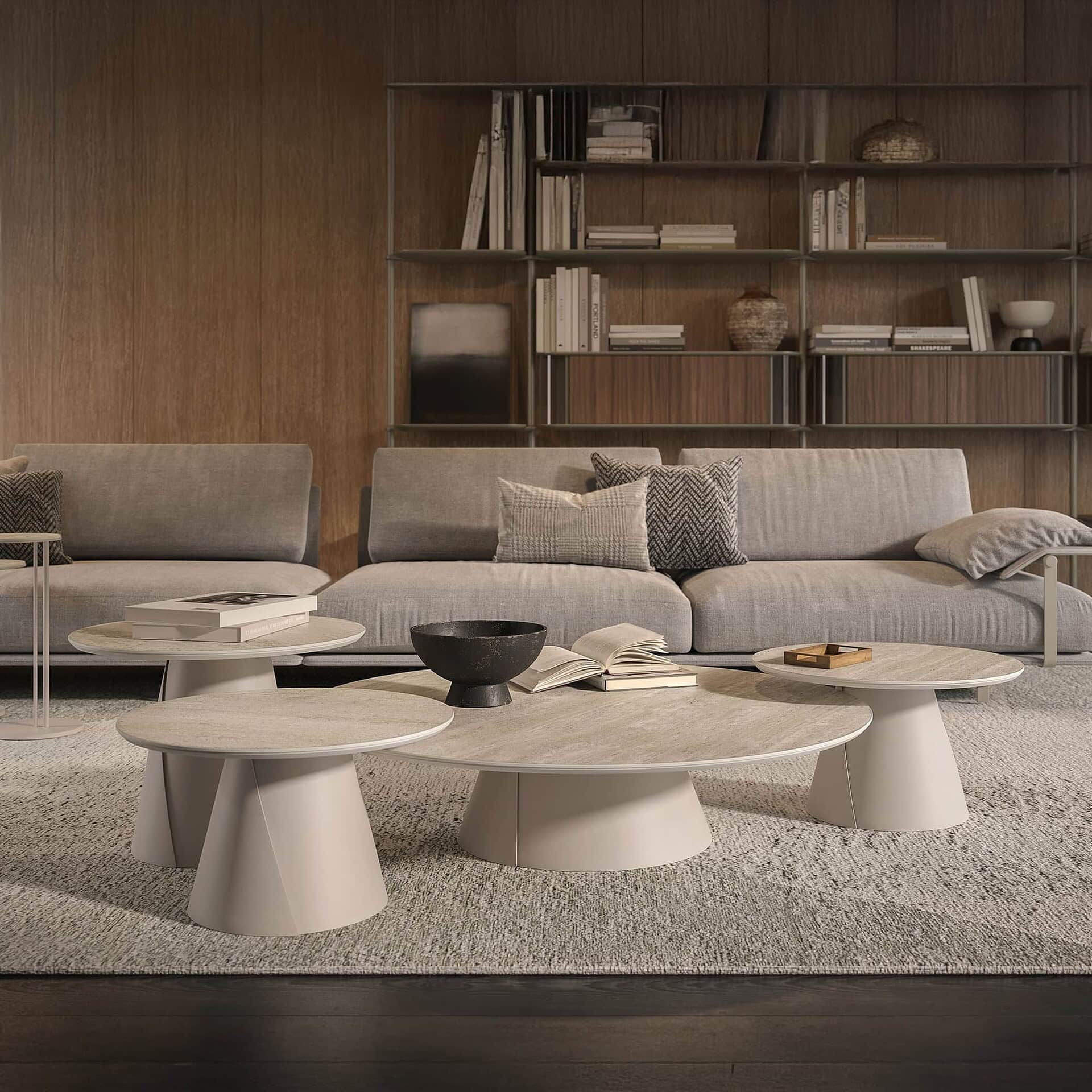 A modern living room with light gray sofas, a cluster of round, light-colored coffee tables, and a wood-paneled wall with a bookshelf filled with books and decor. A textured rug covers the floor.