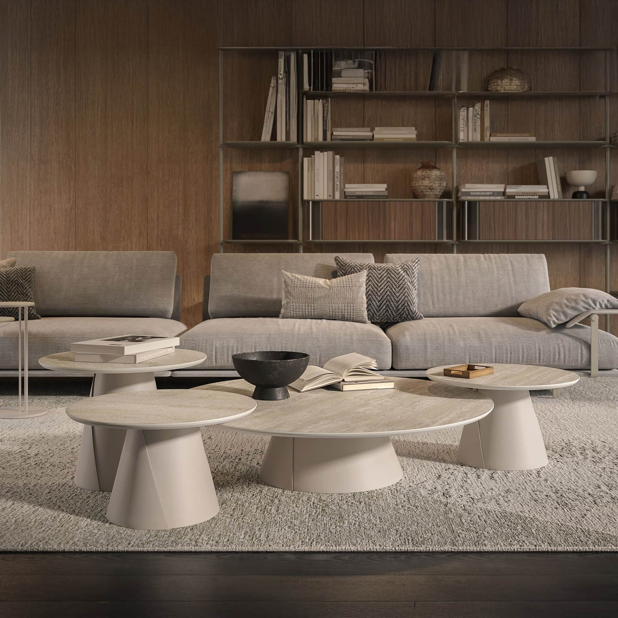 A modern living room with light gray sofas, a cluster of round, light-colored coffee tables, and a wood-paneled wall with a bookshelf filled with books and decor. A textured rug covers the floor.