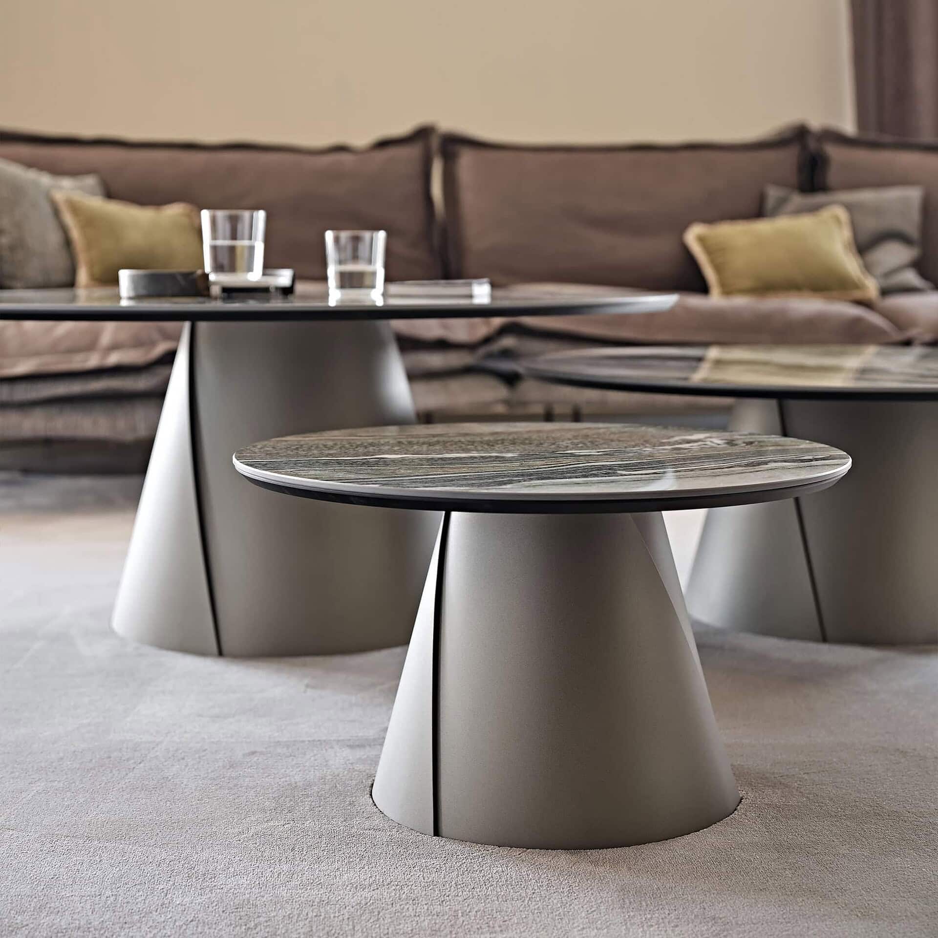 Three modern round coffee tables with veined marble tops and light grey conical bases, arranged in a living room with a plush brown sofa and light grey carpet.