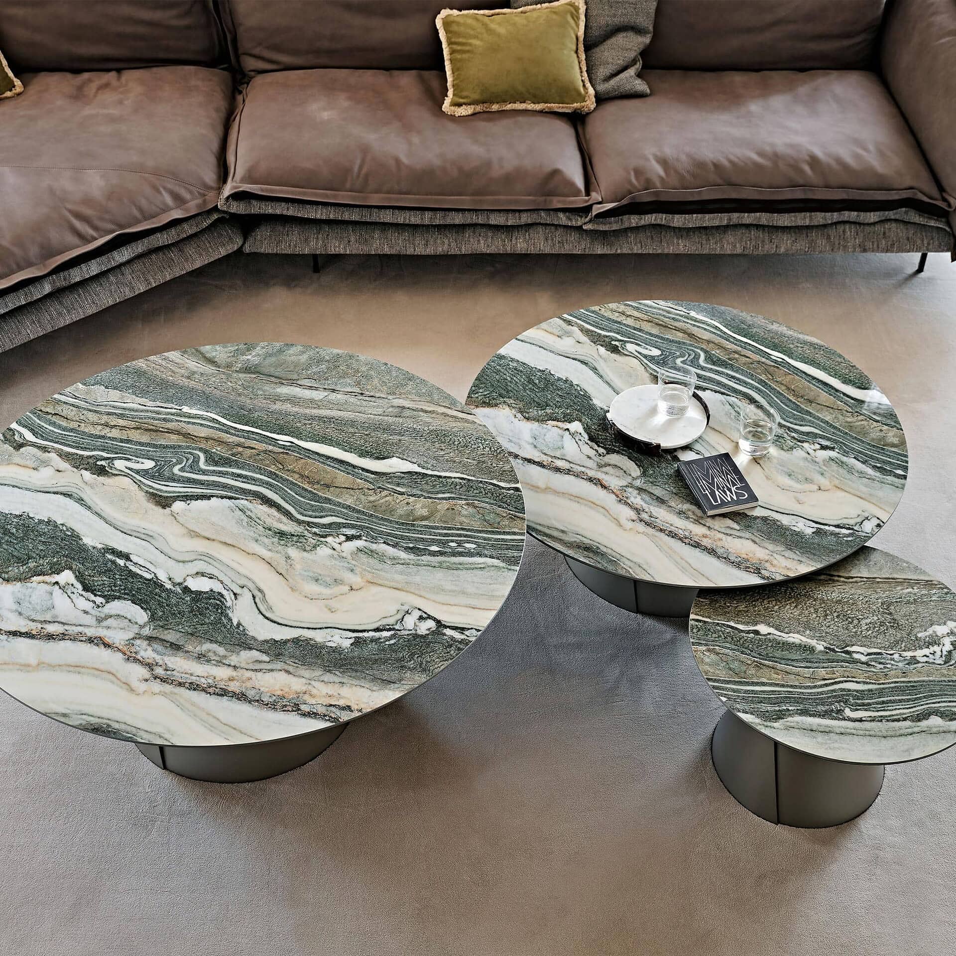 High-angle view of three circular nesting coffee tables with green and white marble tops, in front of a brown leather sectional sofa. Two glasses of water and a book are on the largest table.