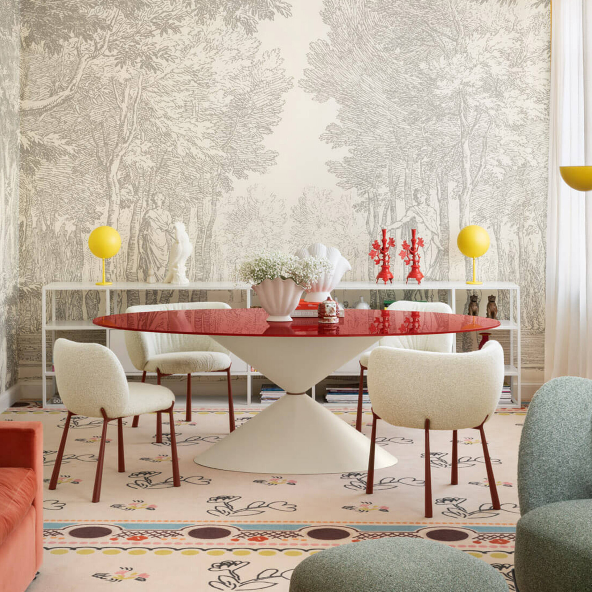 An eclectic dining room with toile de Jouy wallpaper, a red oval table, cream chairs, a colorful patterned rug, and vibrant decorative accents like yellow lamps and red candelabras.