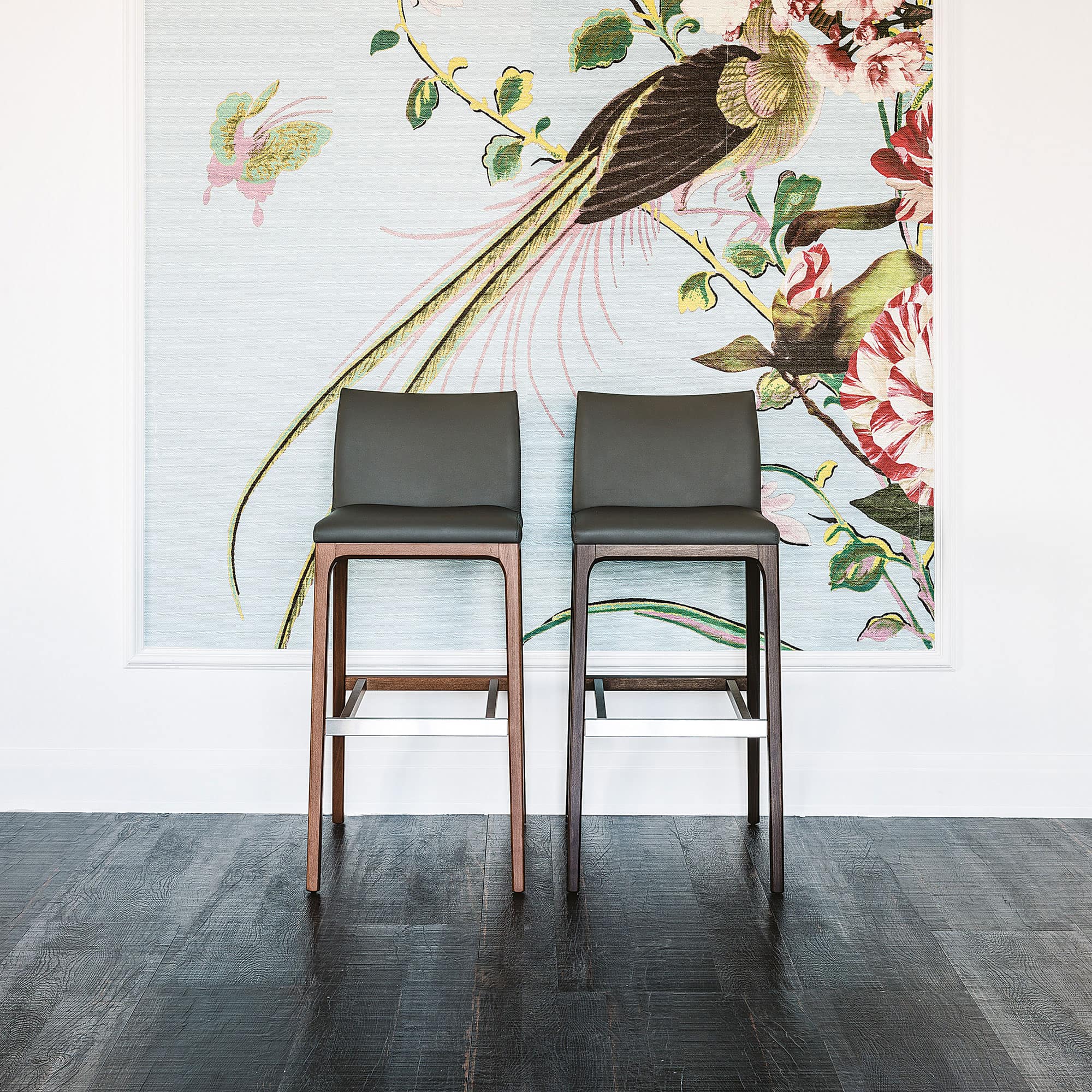Contemporary bar stools with dark green cushions and wooden legs in front of floral wallpaper.
