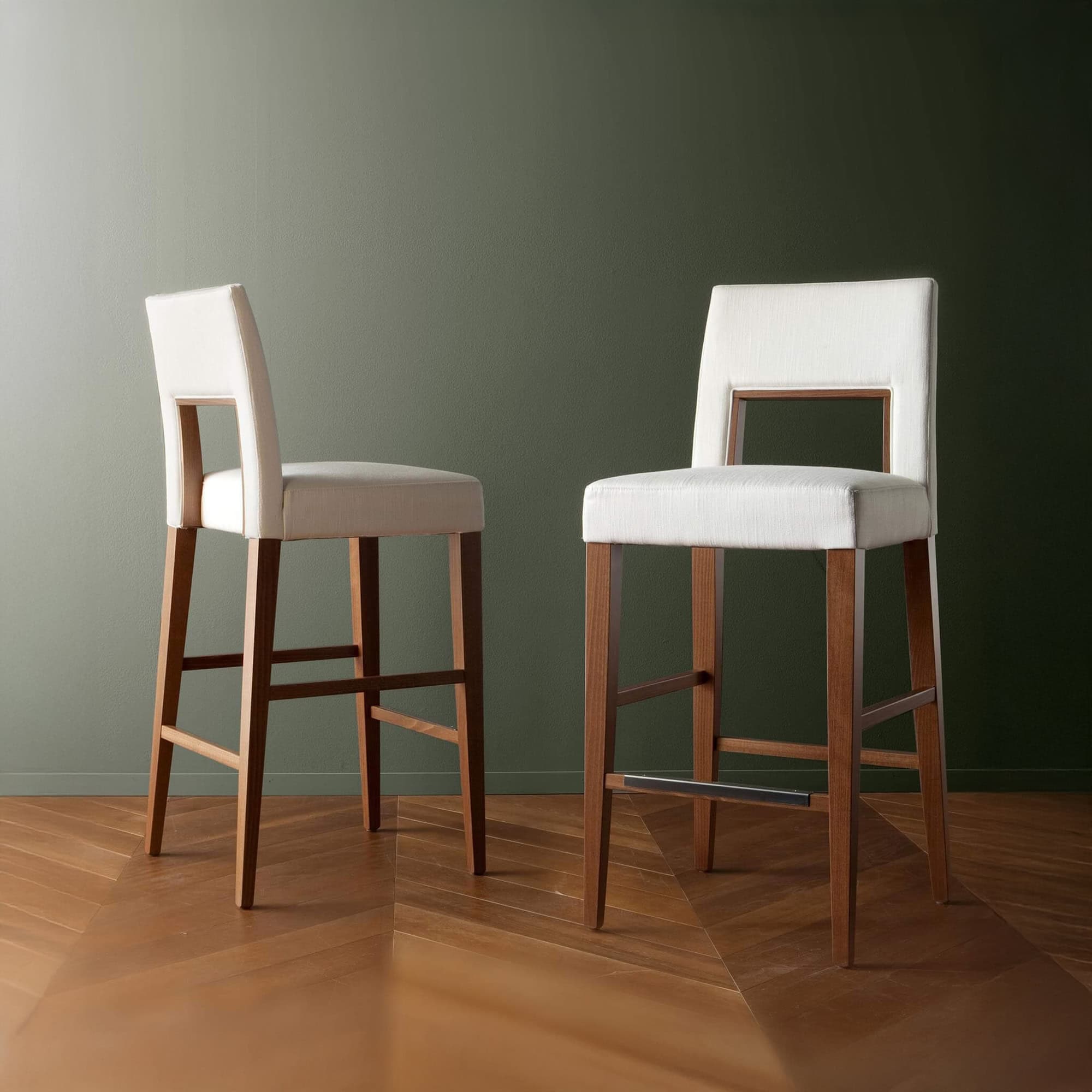 Two modern bar stools with dark wood frames and light cream upholstery, set against a dark olive green wall and a chevron-patterned wooden floor.