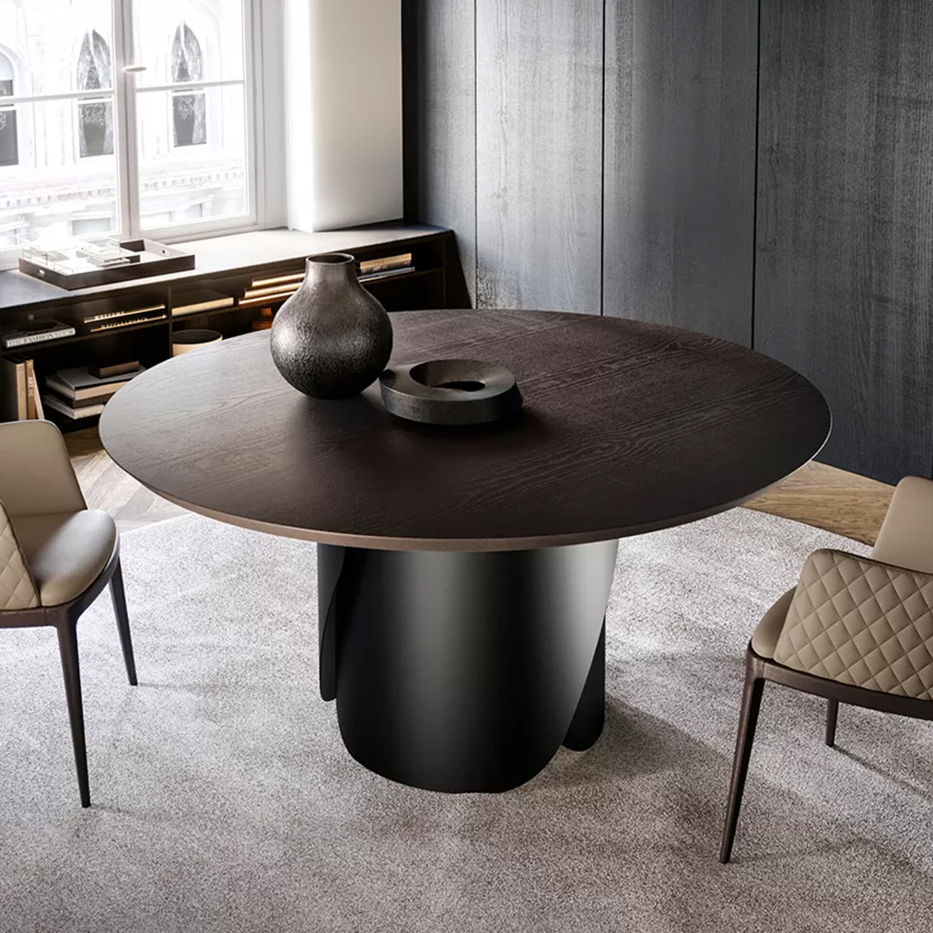 A modern dining room featuring a large dark brown round table with a sculptural black base, two light beige quilted chairs, and a dark paneled wall, illuminated by a large window.