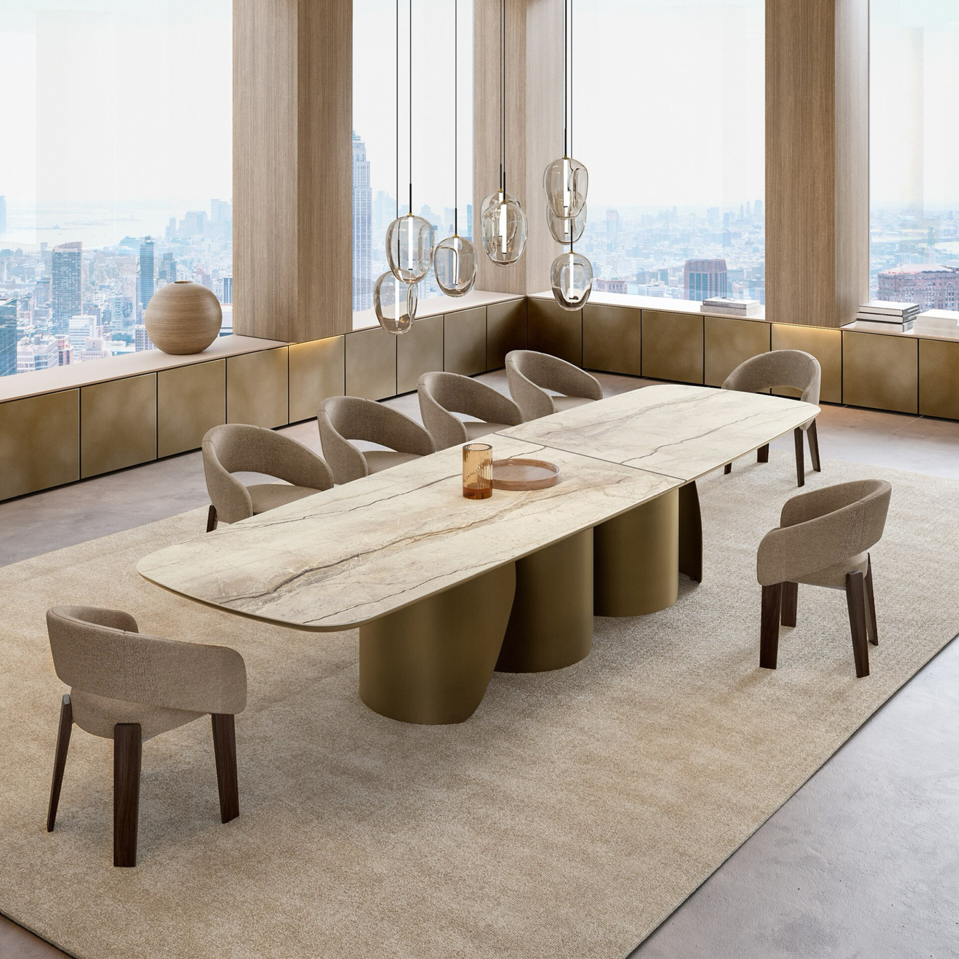 A modern dining room with a large marble-top table, ten beige chairs, and artistic pendant lights, set against a panoramic city skyline view from floor-to-ceiling windows. A low sideboard with a vase and books runs along the window.
