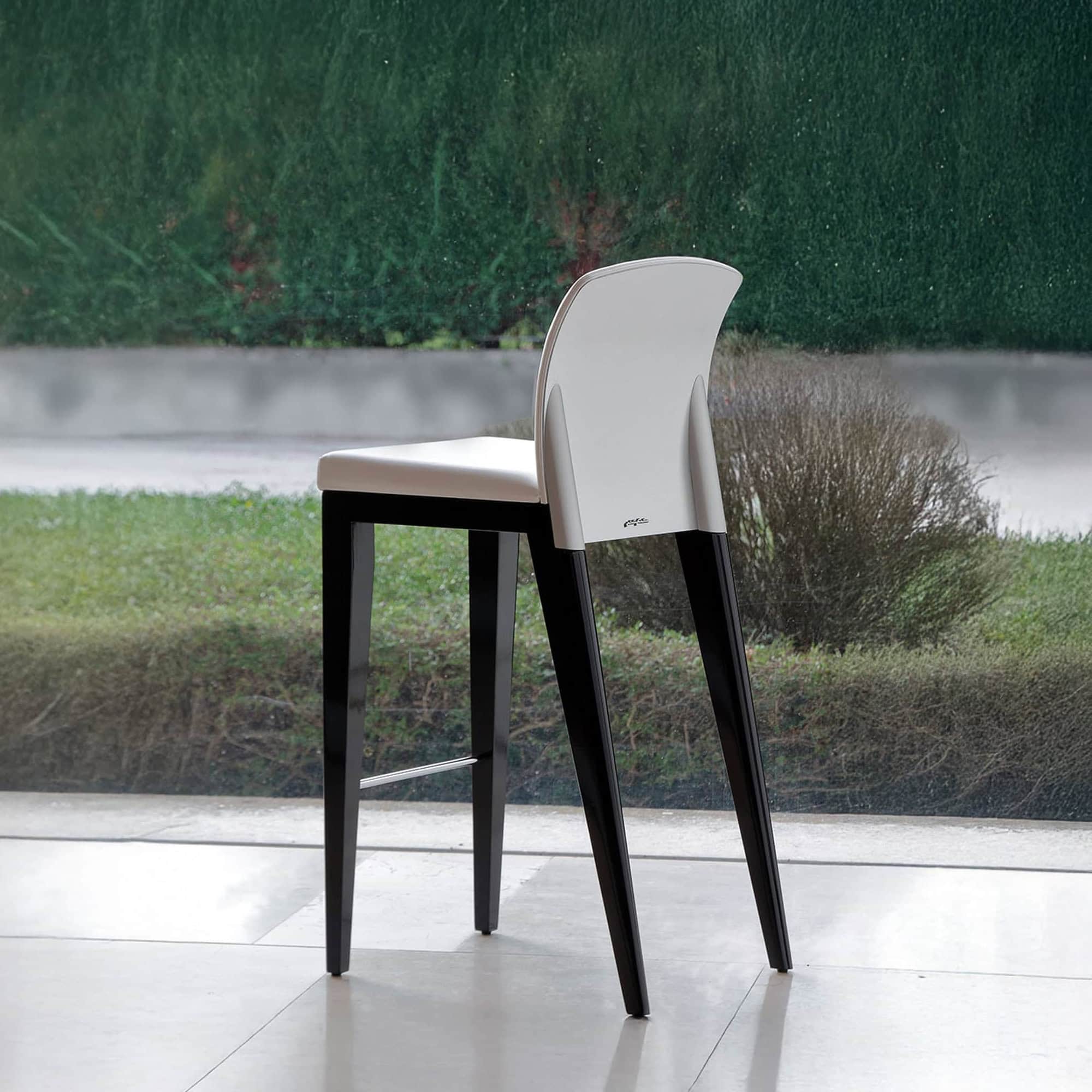 Front angled view of the Sit Bar Stool featuring a white leather seat and black wood legs, placed indoors near large windows.