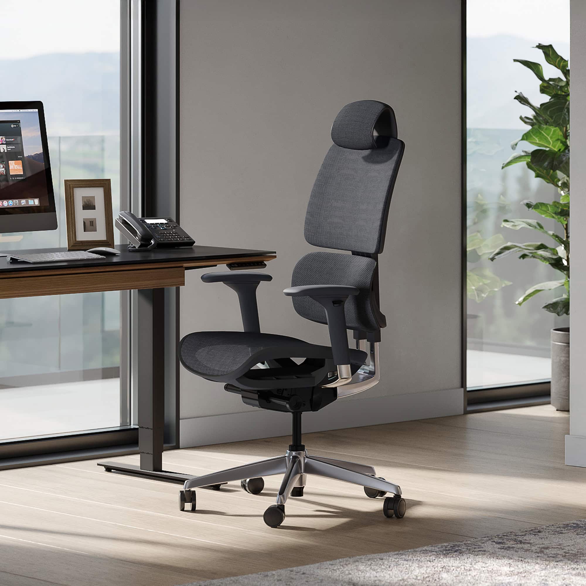 A dark gray ergonomic office chair in a modern home office with a desk, computer, and large window overlooking a landscape.