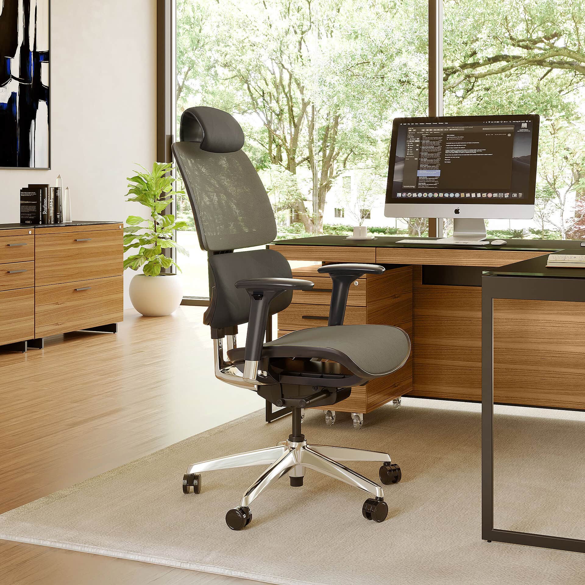 An ergonomic office chair in a modern home office with a wooden desk, iMac computer, and a large window overlooking green trees.