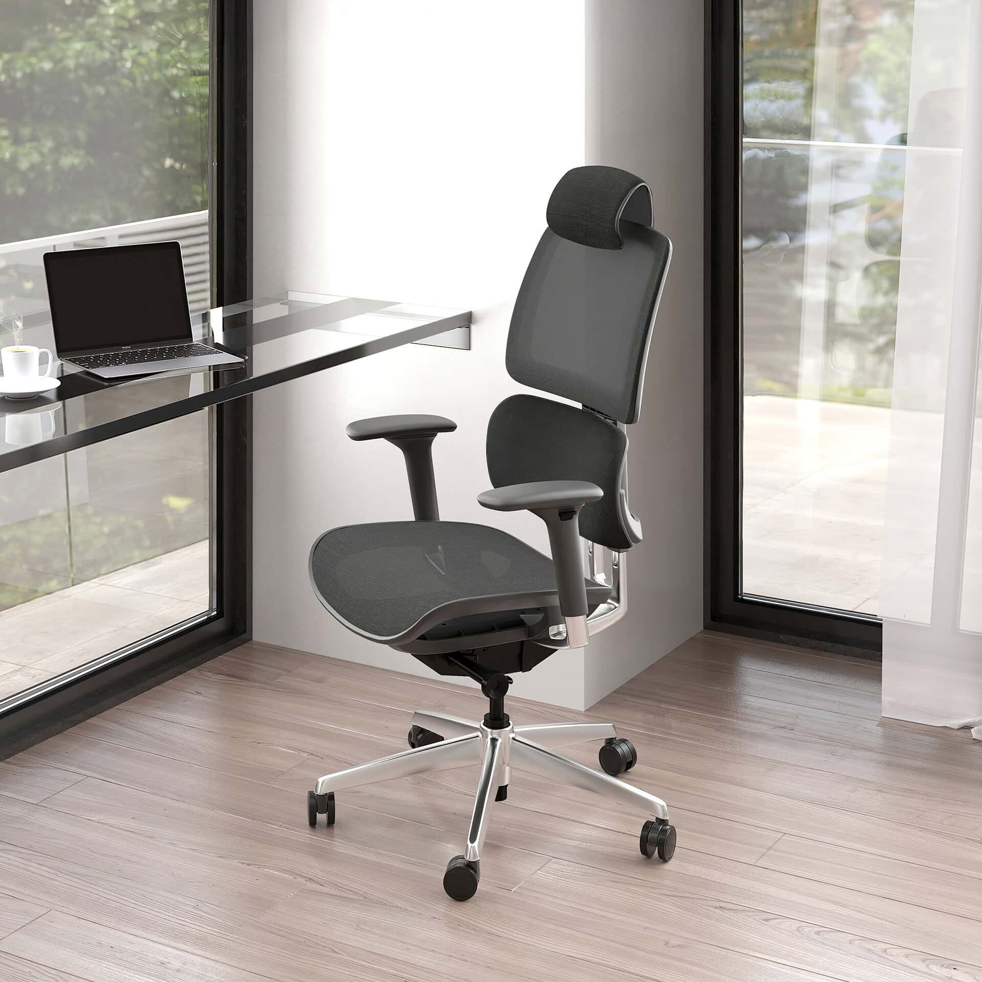 A dark gray ergonomic office chair with a mesh back and headrest, positioned in a bright modern office corner with a black glass desk, laptop, and large windows overlooking green foliage.