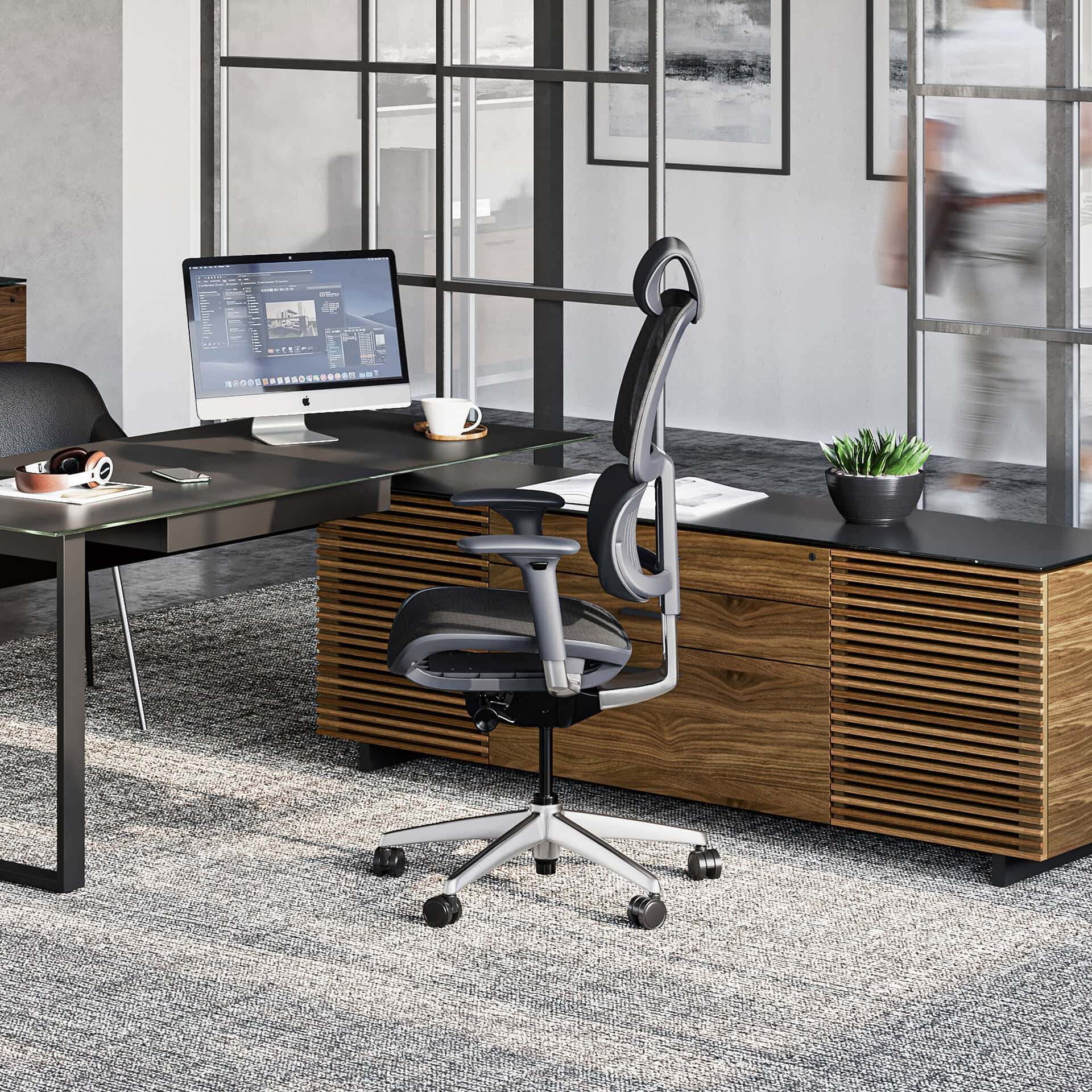 A modern office workspace featuring an L-shaped black desk, an ergonomic grey and black mesh chair, an iMac computer, and a wooden storage unit, set against a glass partition.