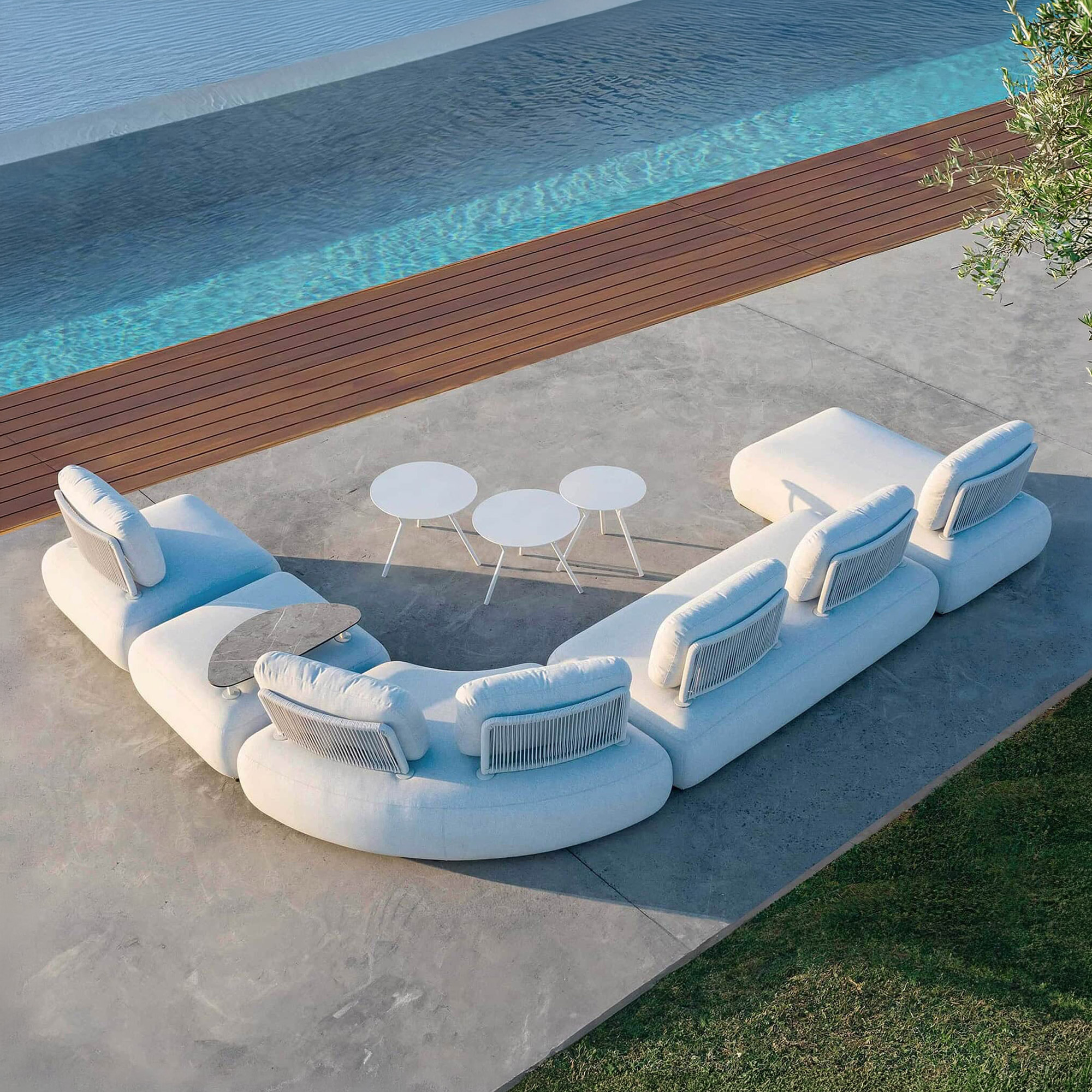 Aerial view of a large, U-shaped modular outdoor sofa in light grey, with three small white side tables, on a concrete patio next to a blue swimming pool with a wooden deck and green grass.