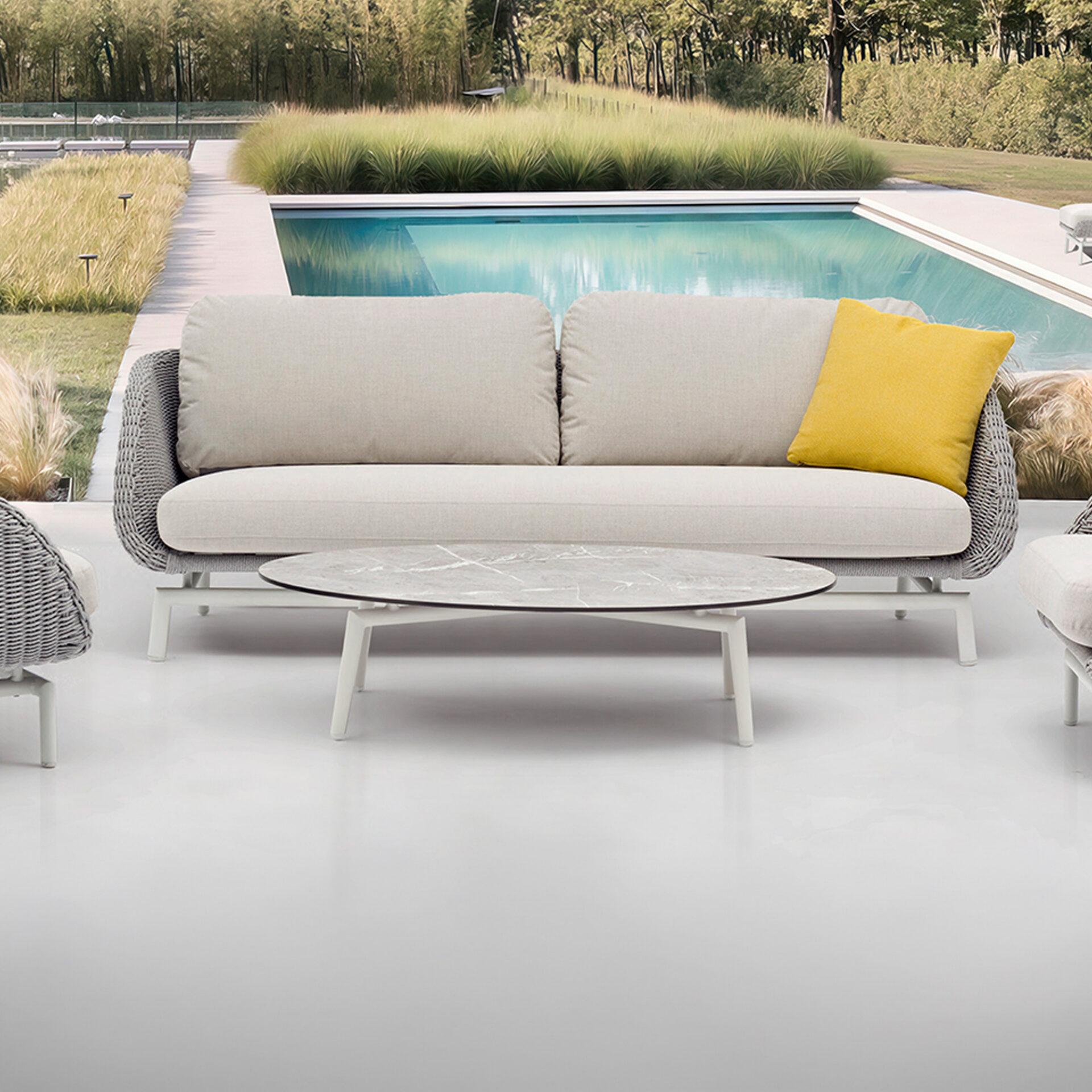 A modern outdoor sofa with beige cushions and a grey woven frame, an oval marble-top coffee table, and a yellow throw pillow on a light grey patio next to a blue swimming pool with lush green and golden grasses in the background.
