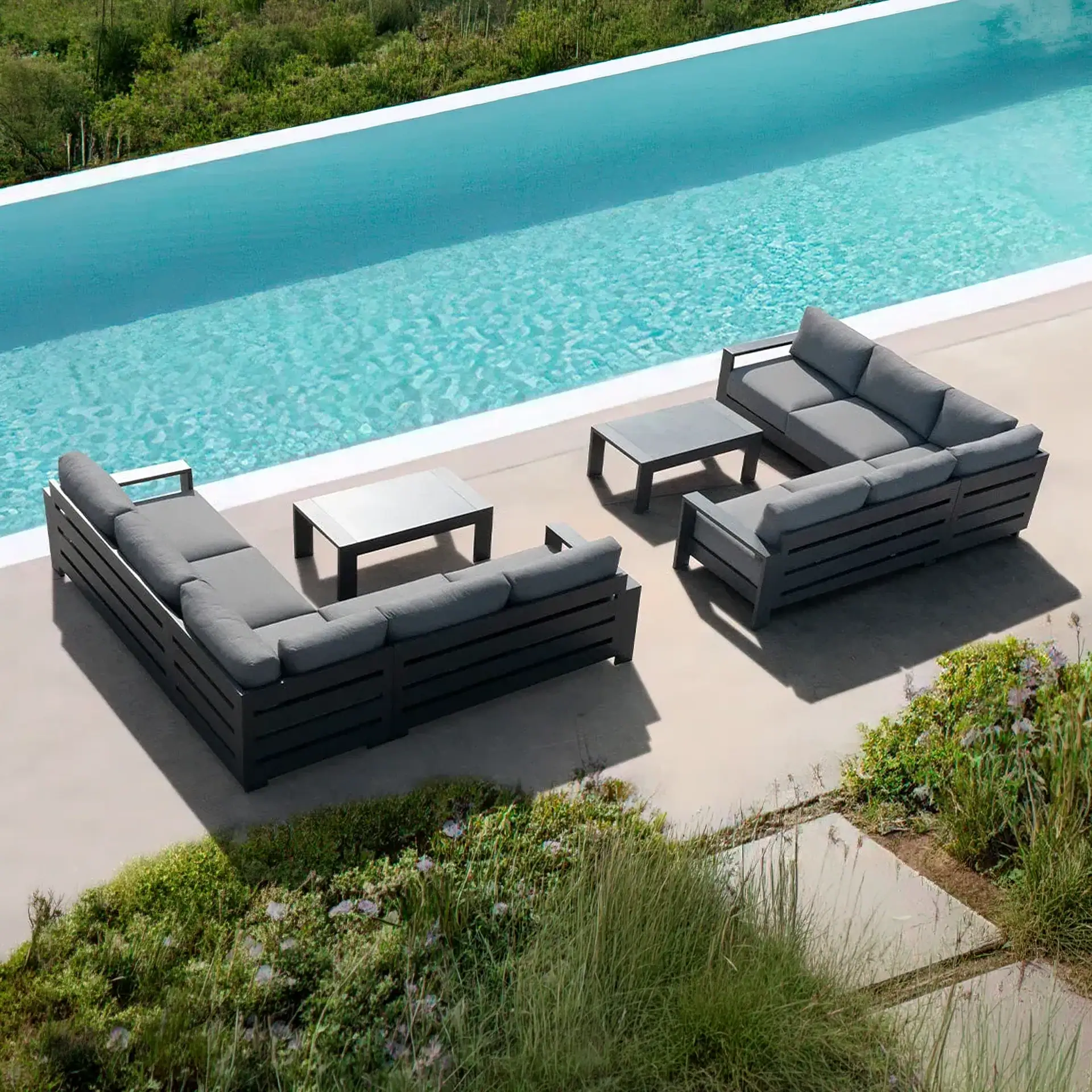An outdoor patio with dark gray sectional sofas, light gray cushions, and coffee tables beside a blue swimming pool, surrounded by green foliage.