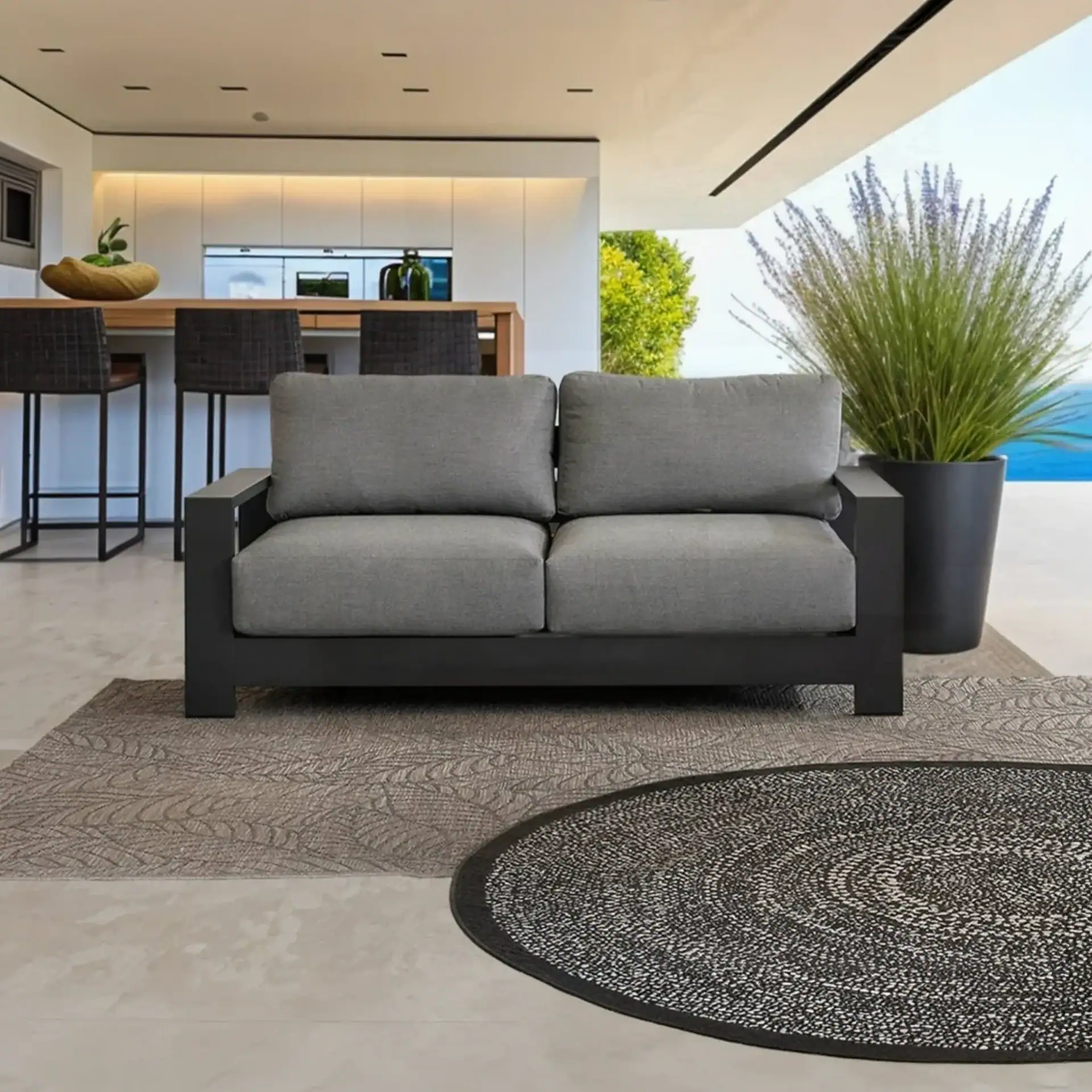 A grey two-seater outdoor sofa with a dark frame sits on a patterned rug. In the background is a modern kitchen and a potted plant with a view of the ocean.